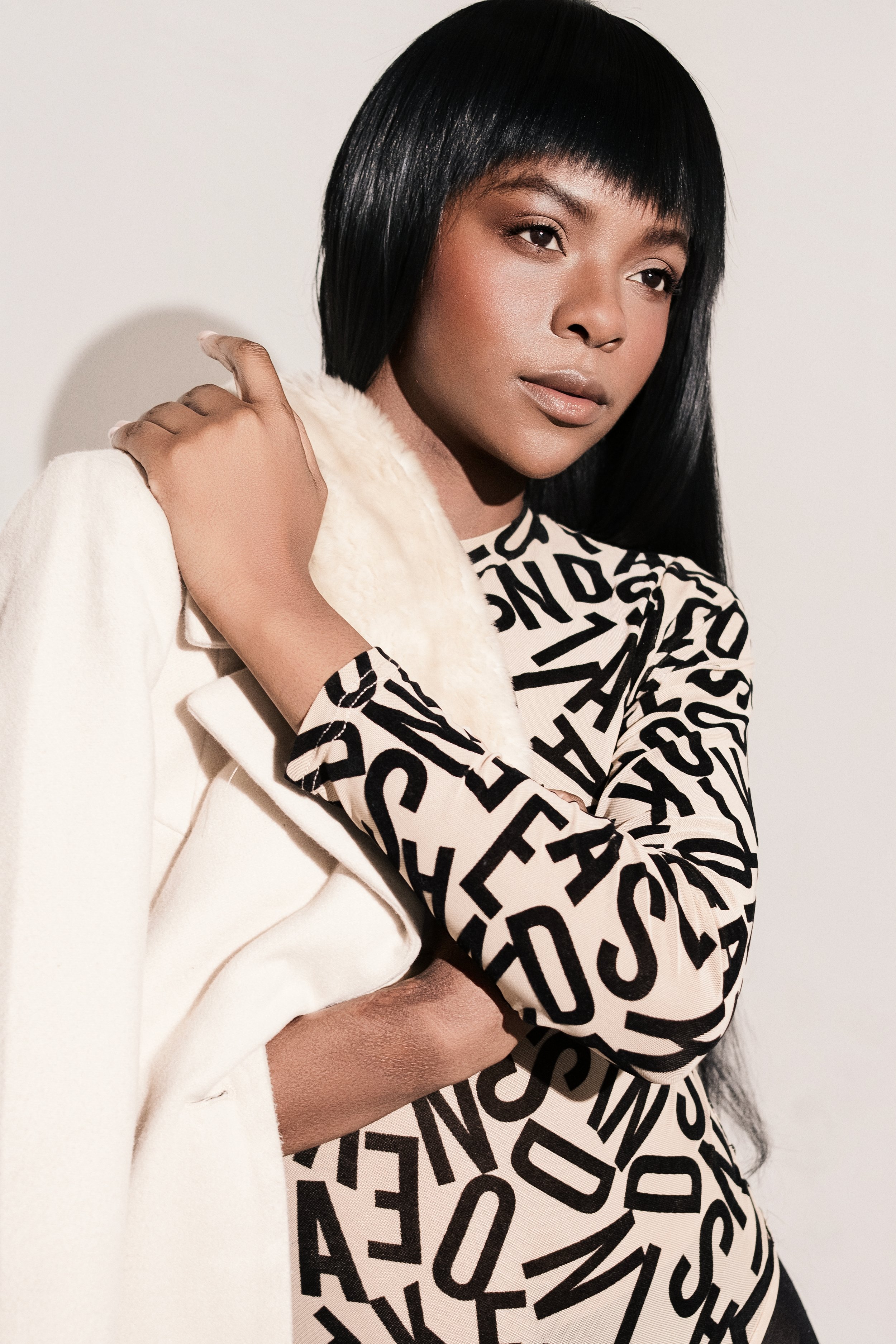 Woman with black hair in a bob cut, wearing a long sleeve shirt with black letters, holding a cream-colored coat or scarf.