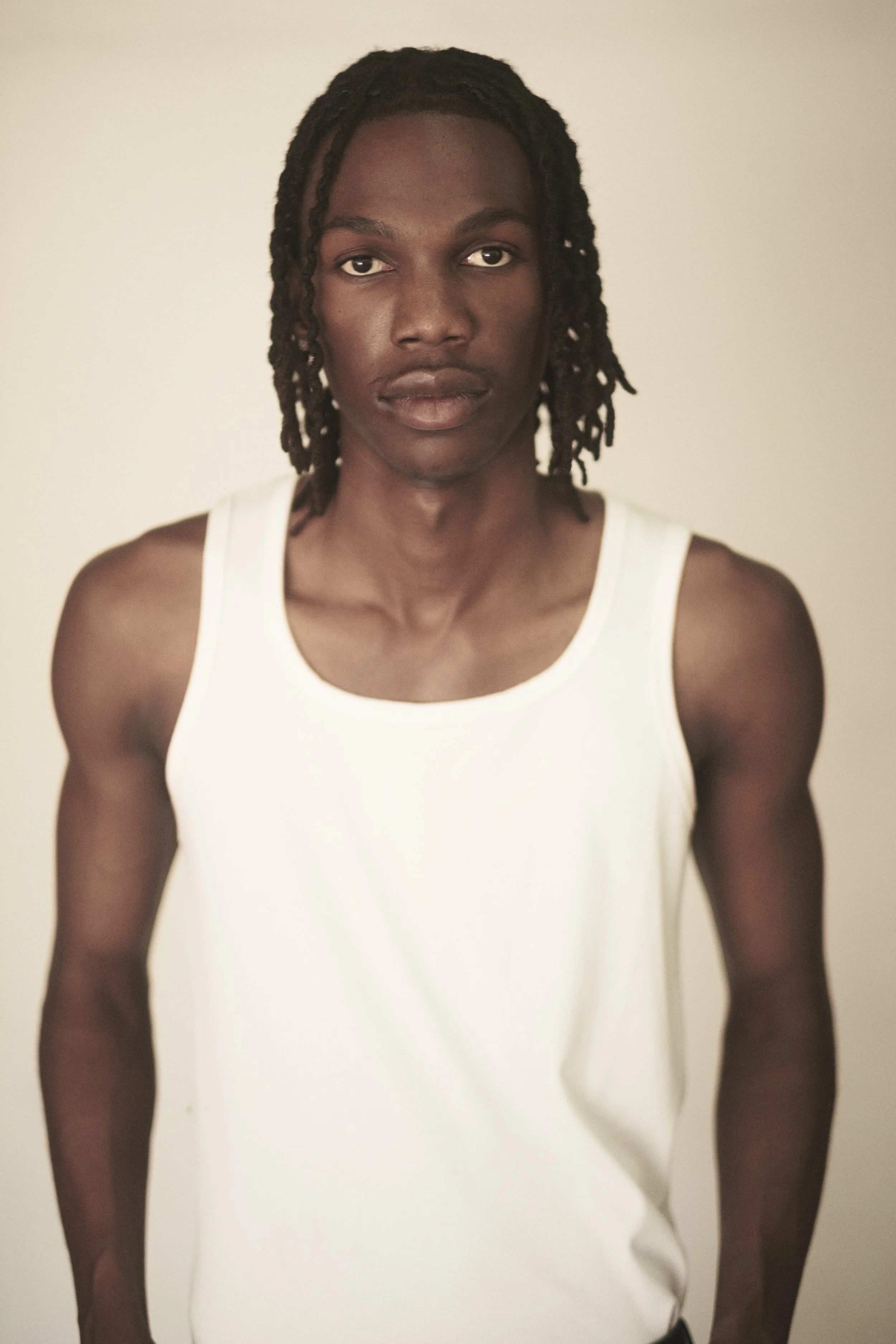 TSHEDZA Ramavheya from AURA Models with dark skin, dreadlocked hair, wearing a white tank top, standing against a plain light-colored background.