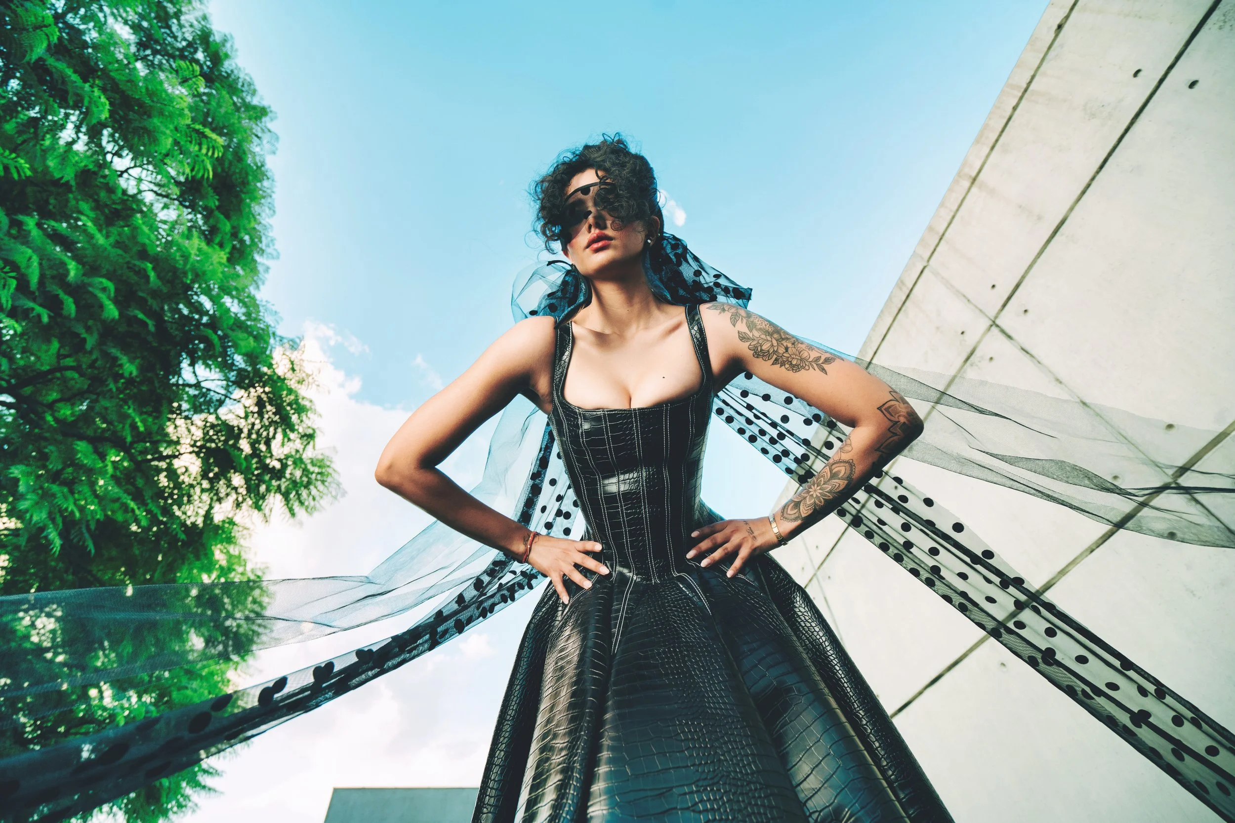 EMILY Francis from AURA Models wearing a black dress and a sheer, polka dot cape standing outdoors under a clear blue sky, with green trees and a concrete structure nearby.