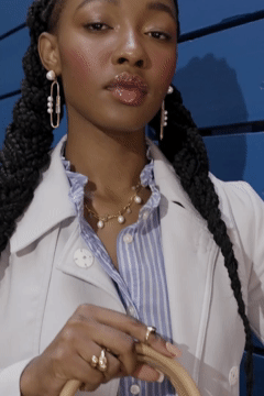 A woman with braided hair wearing pearl earrings, a pearl necklace, a striped shirt, and a white coat, holding a bag.