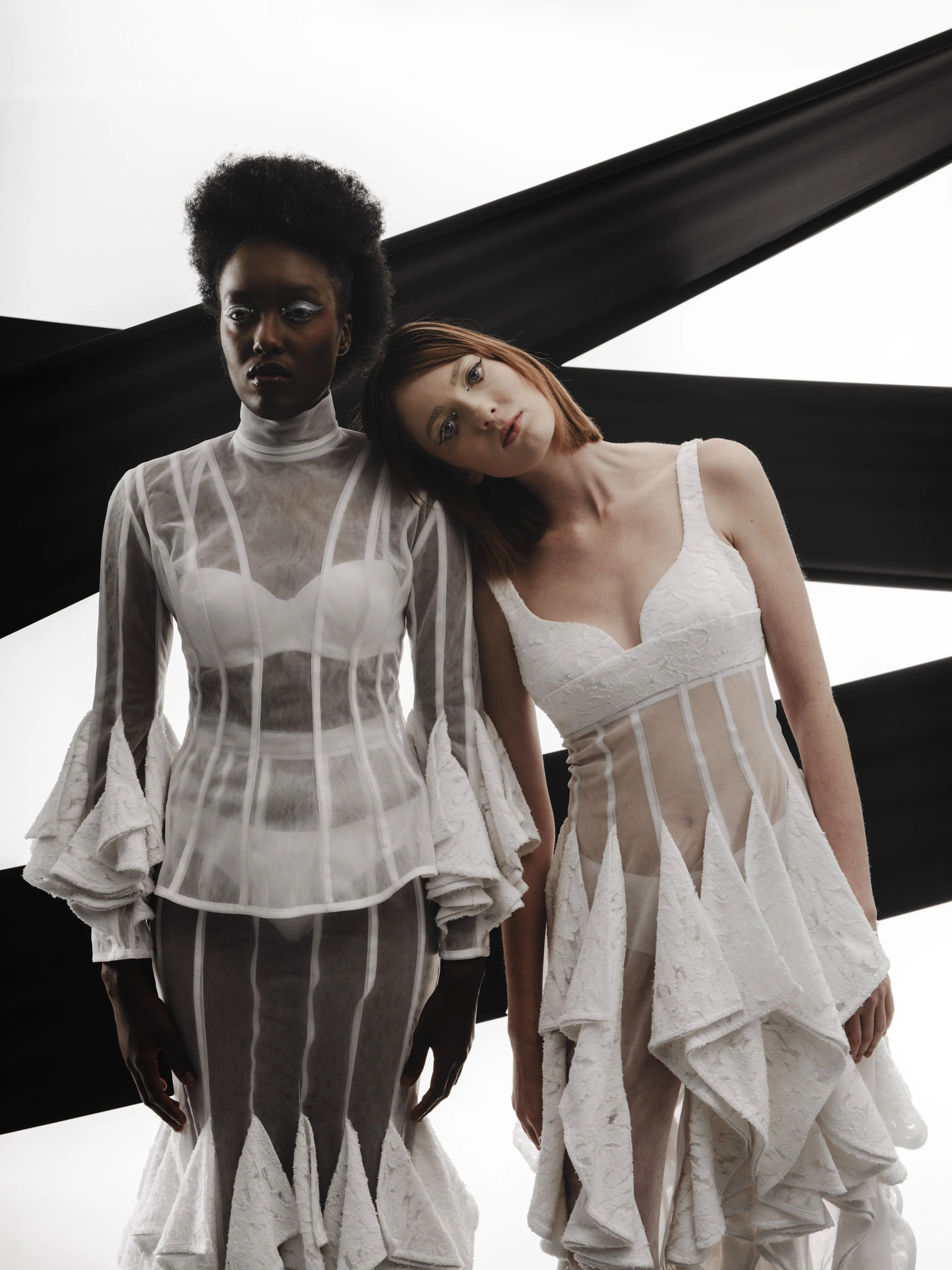 CAYLEIGH Bancroft and KIM Chakanyuka from AURA Models standing close together, wearing white garments with layered, textured styles, set against a stark white background with black beams.