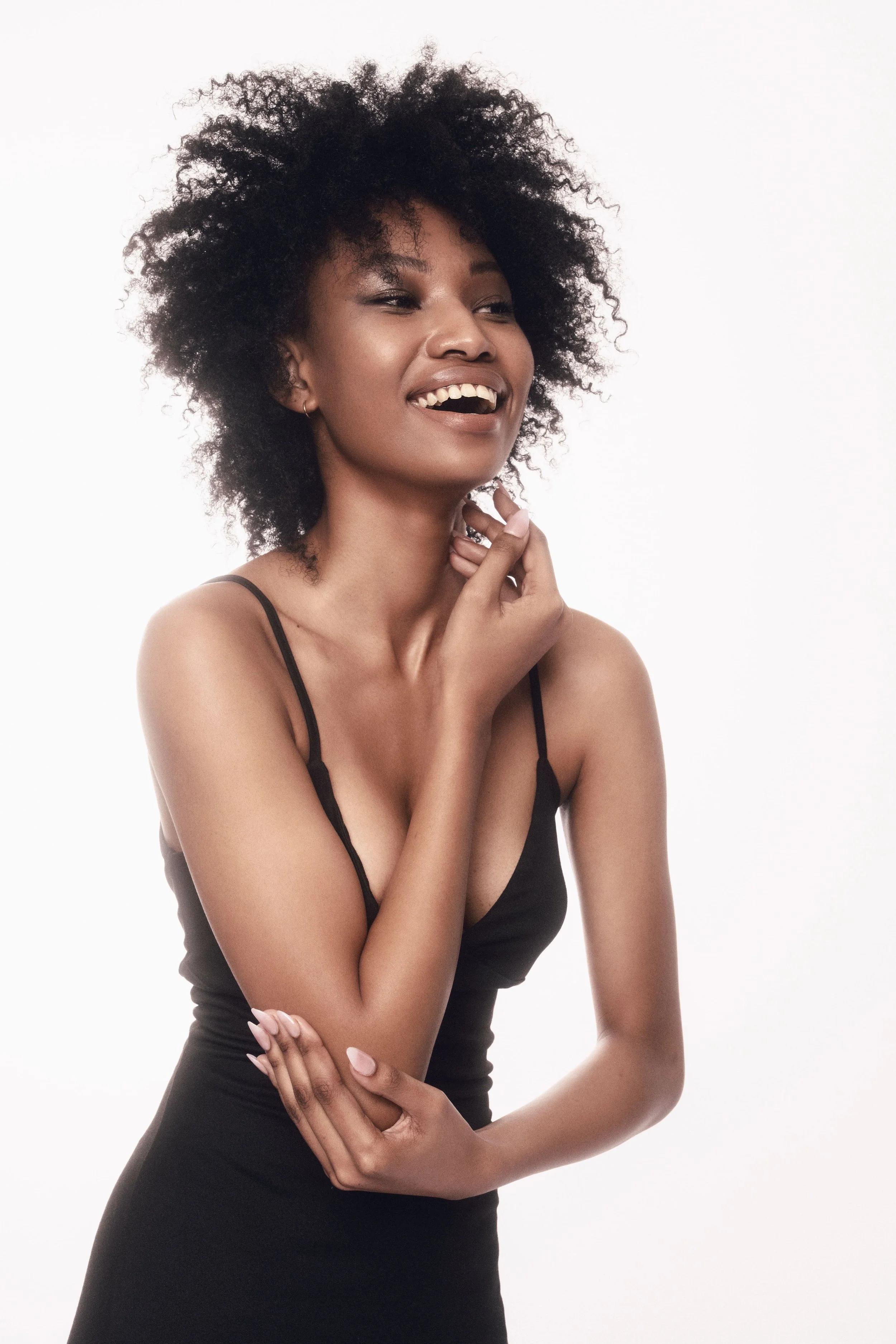 QUISHA Rapoo from AURA Models with dark, curly hair and light brown skin, wearing a black spaghetti strap dress, posing against a plain white background, smiling, with her right hand touching her face and her left arm crossed over her stomach.