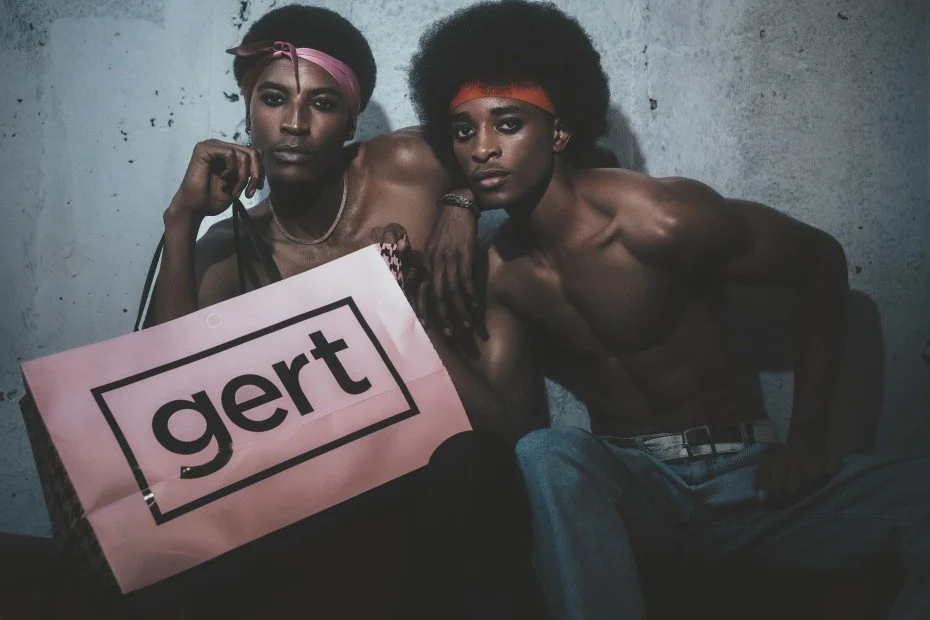 Two individuals with dark skin and wearing headbands, KUNDAI Madhandara from AURA Models in pink and the other one orange, pose shirtless against a plain wall. They hold a pink shopping bag with the word "gert" printed on it.