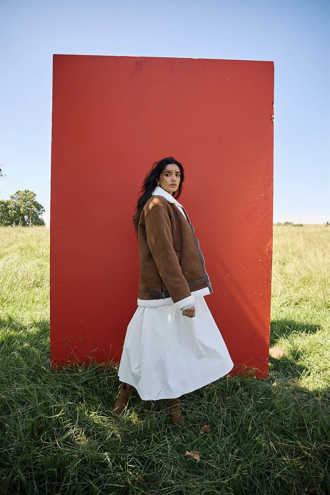 EMILY Francis from AURA Models standing outdoors in a grassy field with a large red square behind her. She is wearing a brown jacket with a white fuzzy lining and a long white skirt.