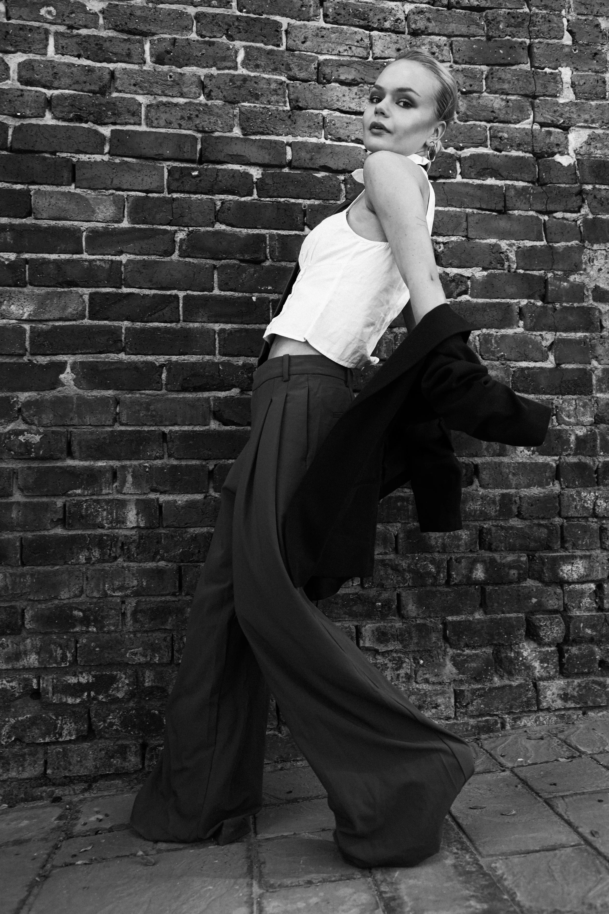 Black and white photo of a woman with platinum blonde hair styled sleekly, leaning against a brick wall. She is dressed in a sleeveless top, relaxed pants, and a blazer draped over her arm, with earrings and makeup highlighting her features.