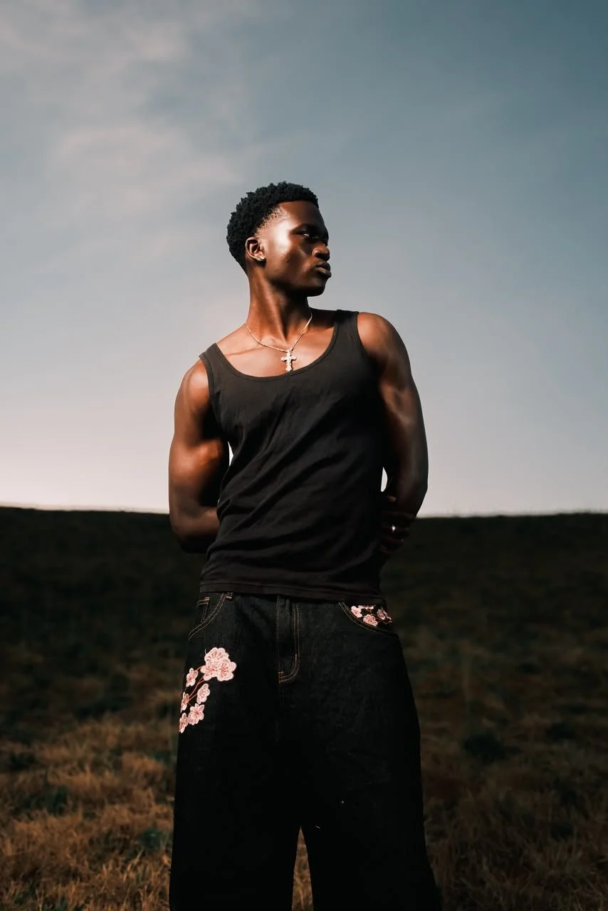 MBHURI Baloyi from AURA Models stands outdoors on a grassy field during sunset, wearing a black tank top, black jeans with floral embroidery, and a cross necklace, with arms behind her back and looking to the side.