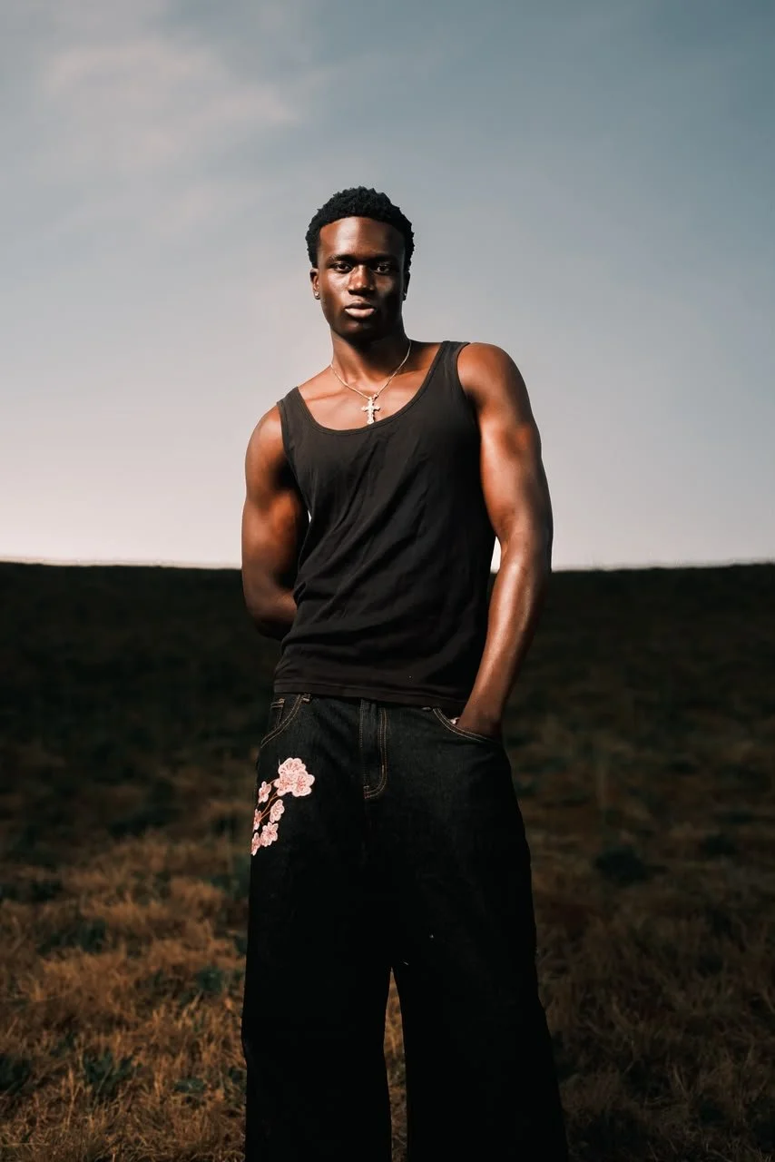 MBHURI Baloyi from AURA Models with short curly hair stands outdoors in a natural landscape at dusk, wearing a black sleeveless top, embroidered black pants, a silver cross necklace, with hands in pockets, looking directly at the camera.