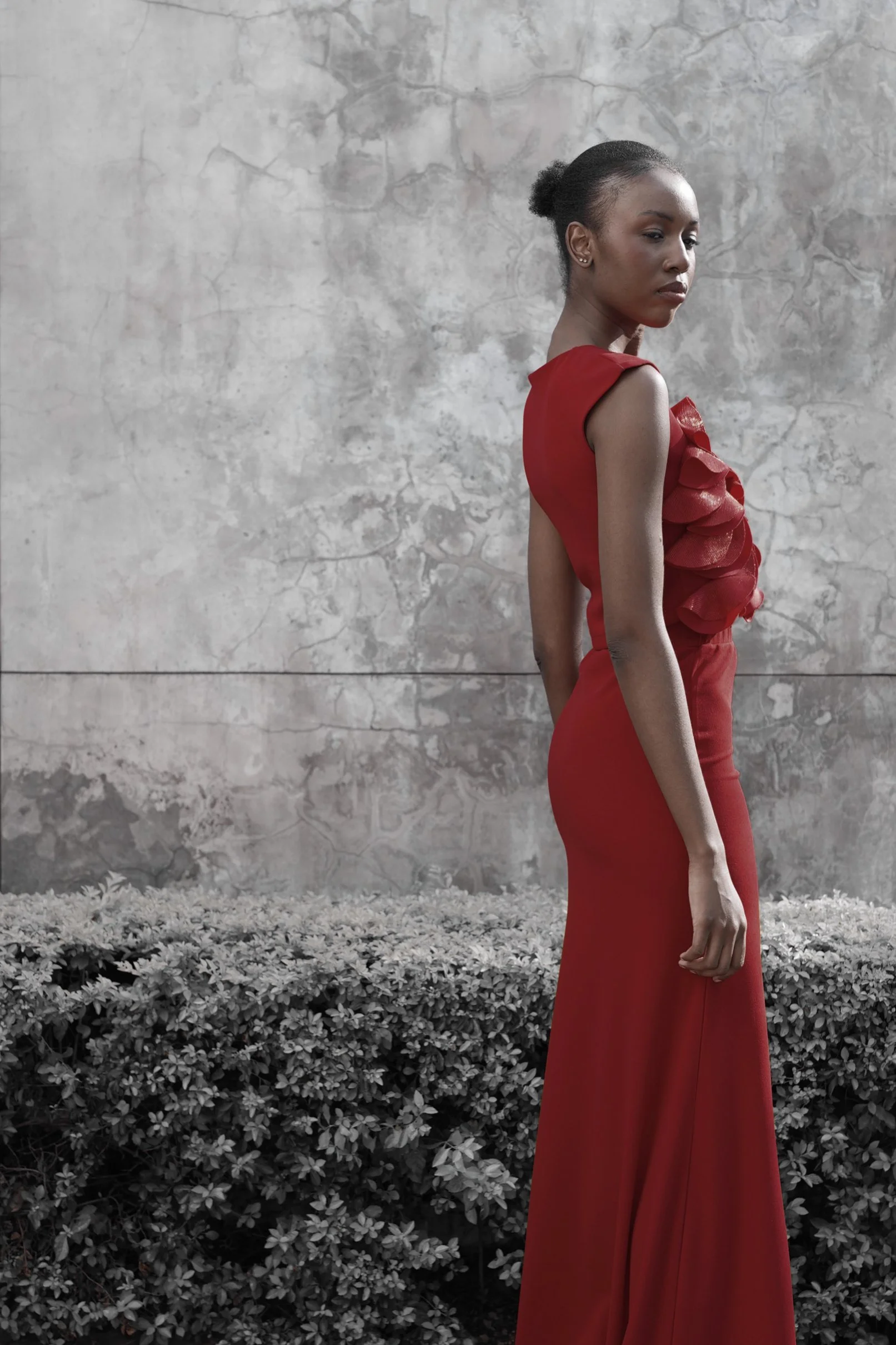 A woman in a red gown standing outdoors against a textured gray stone wall.