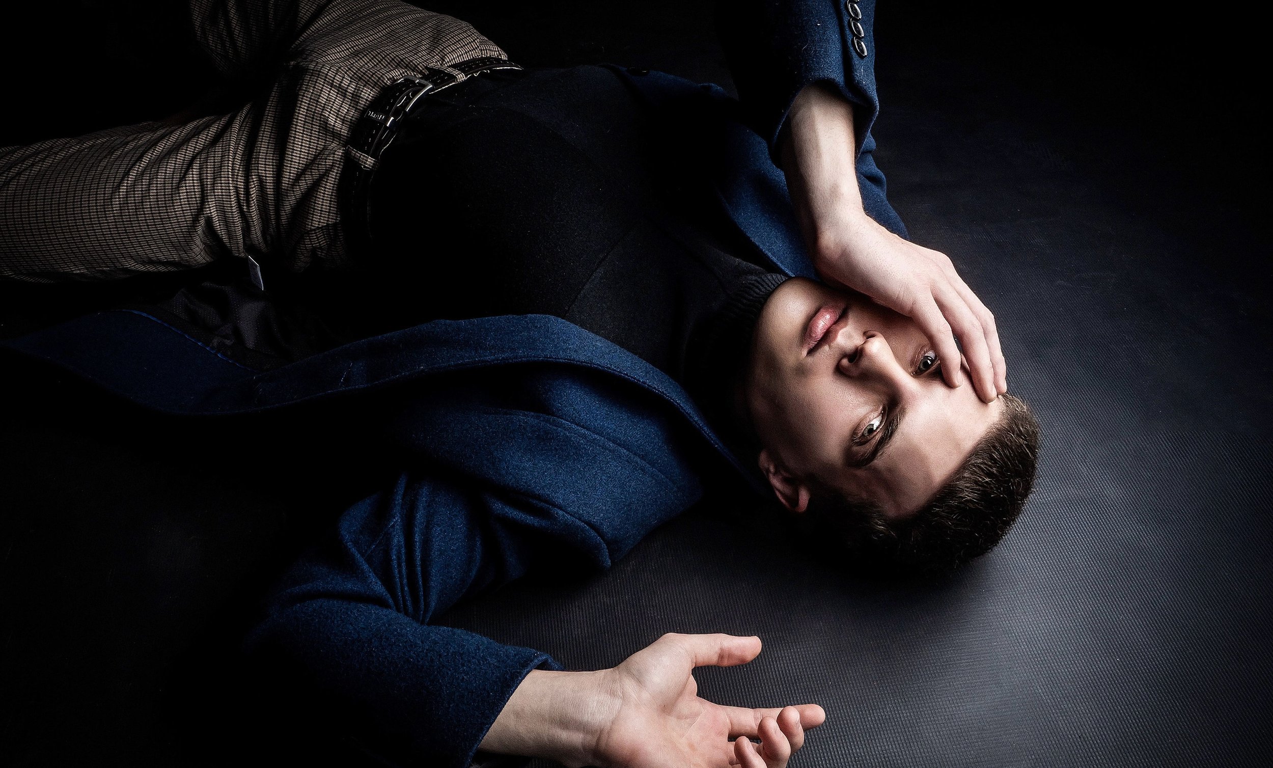 A young man lying on the floor with one hand covering part of his face and the other arm extended, wearing a blue jacket and black shirt, with a dark background.