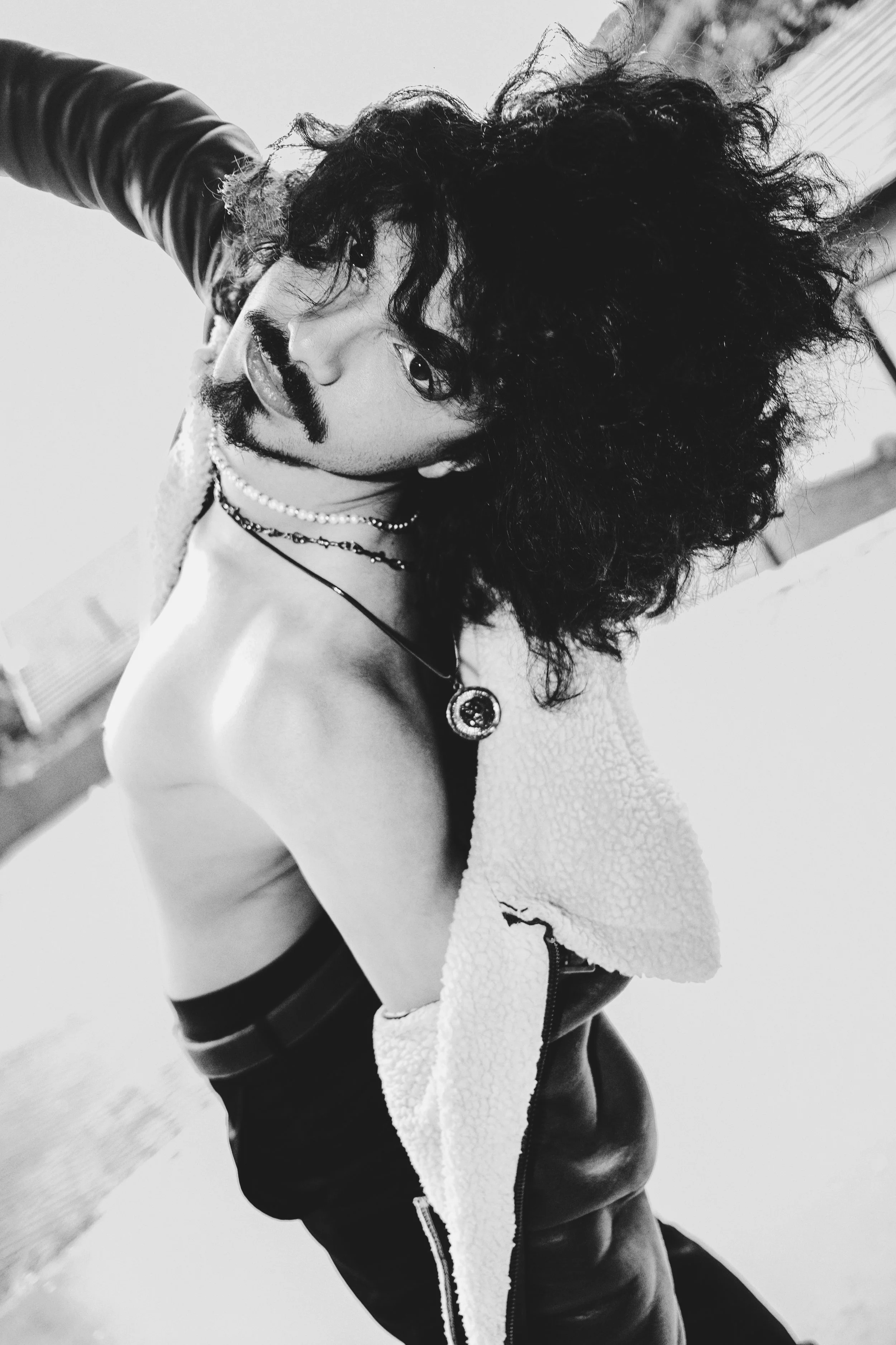 A black and white photo of a person with curly hair, wearing a necklace, a leather jacket, and leather pants, striking a pose with one arm raised and looking directly at the camera.