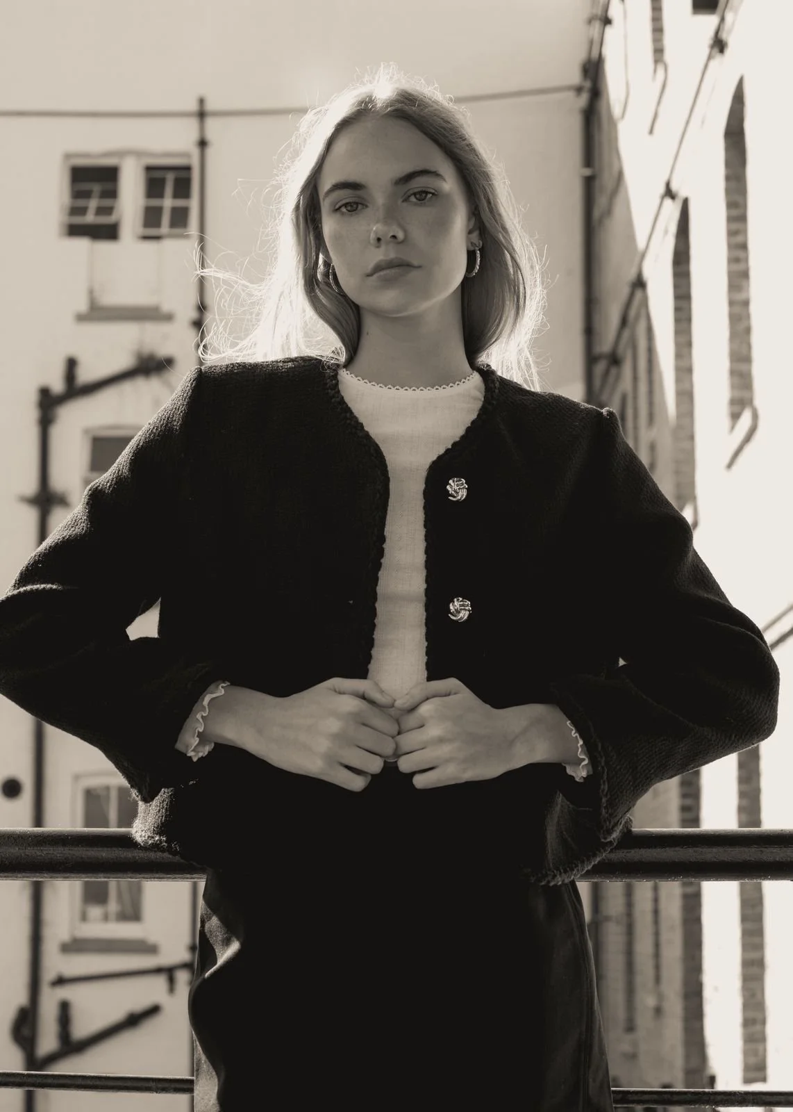 Black and white photo of KEELY Miller from AURA Models with long hair, standing outdoors between buildings, wearing a black jacket with decorative buttons, earrings, and a white top.