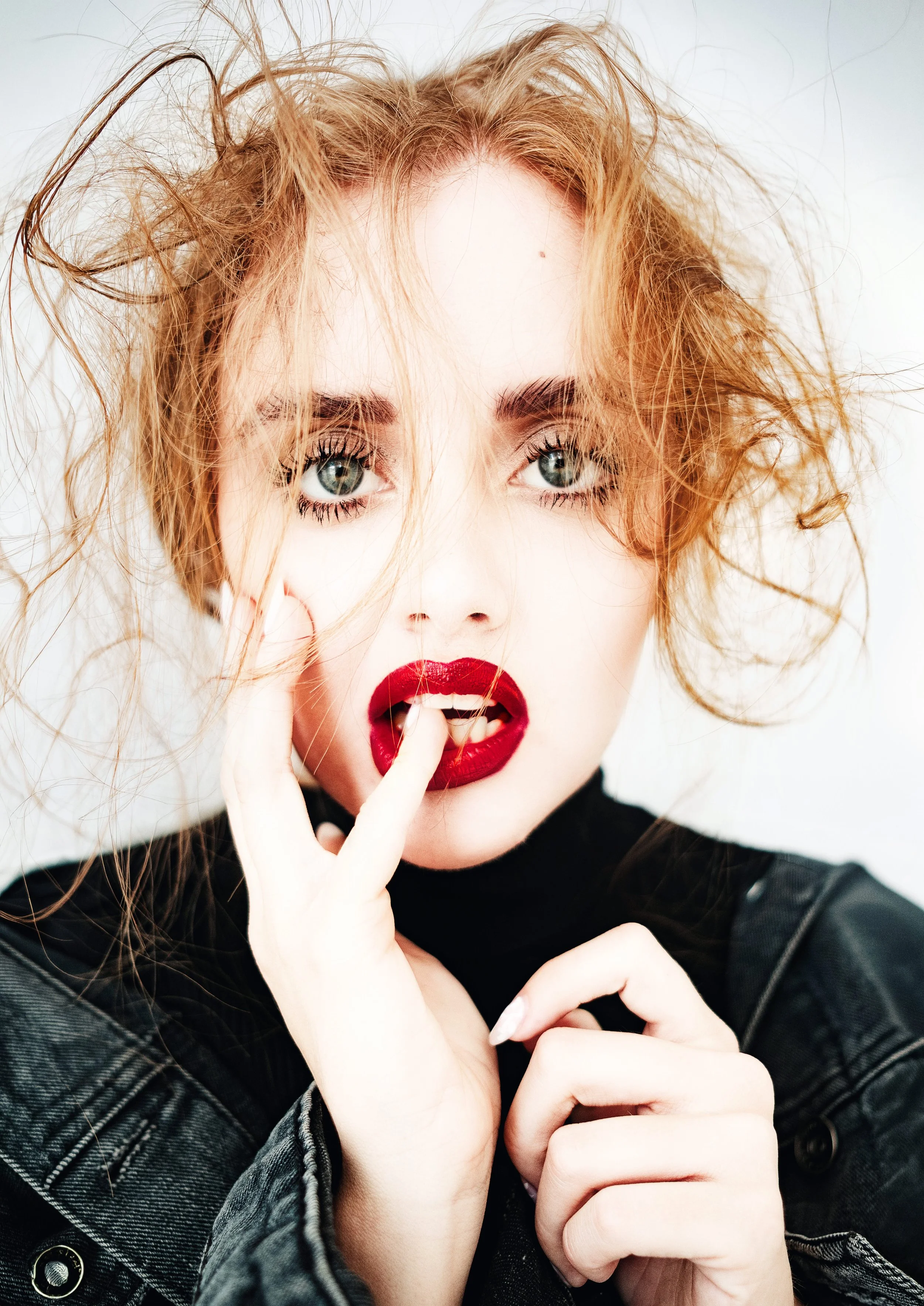 DANIELLE Laubscher from AURA Models with curly red hair, bright blue eyes, wearing red lipstick, biting her finger, and wearing a black leather jacket.
