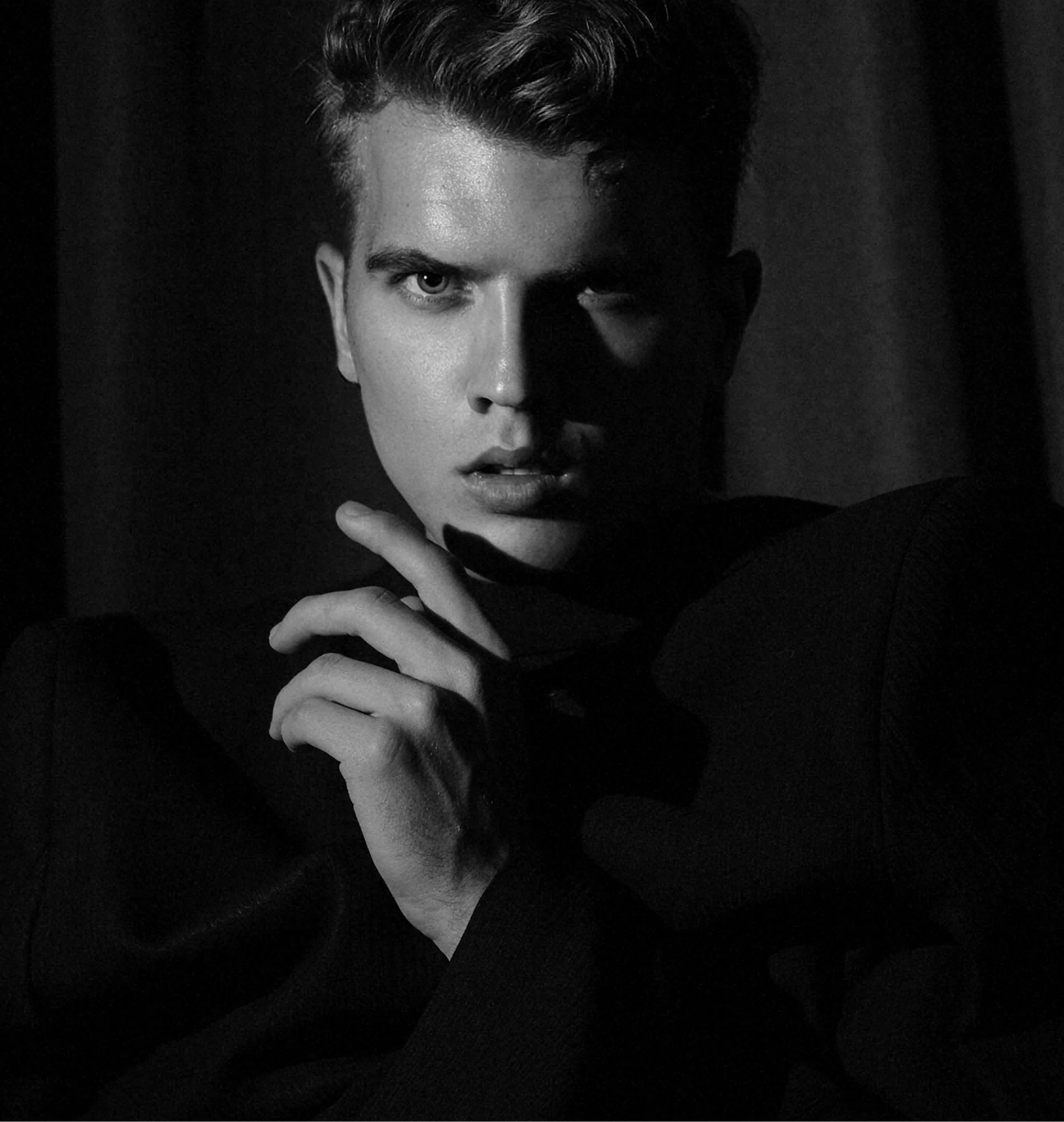 Black and white close-up photo of a young man with styled hair, intense gaze, and partly illuminated face, with hand near chin, wearing a dark jacket.
