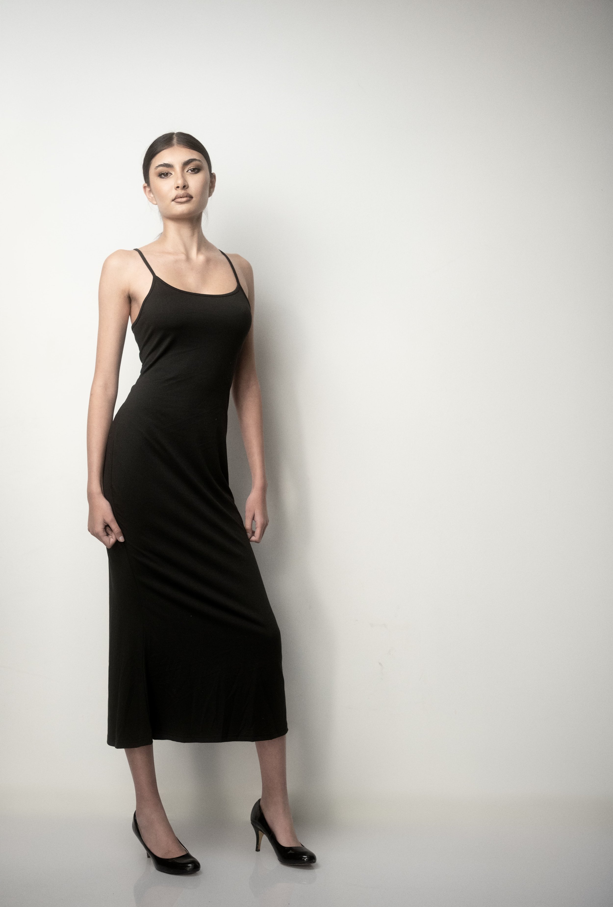 KEIRA Louw from AURA Models with dark hair in a low bun wearing a fitted black spaghetti strap dress and black high heels standing against a plain white wall.