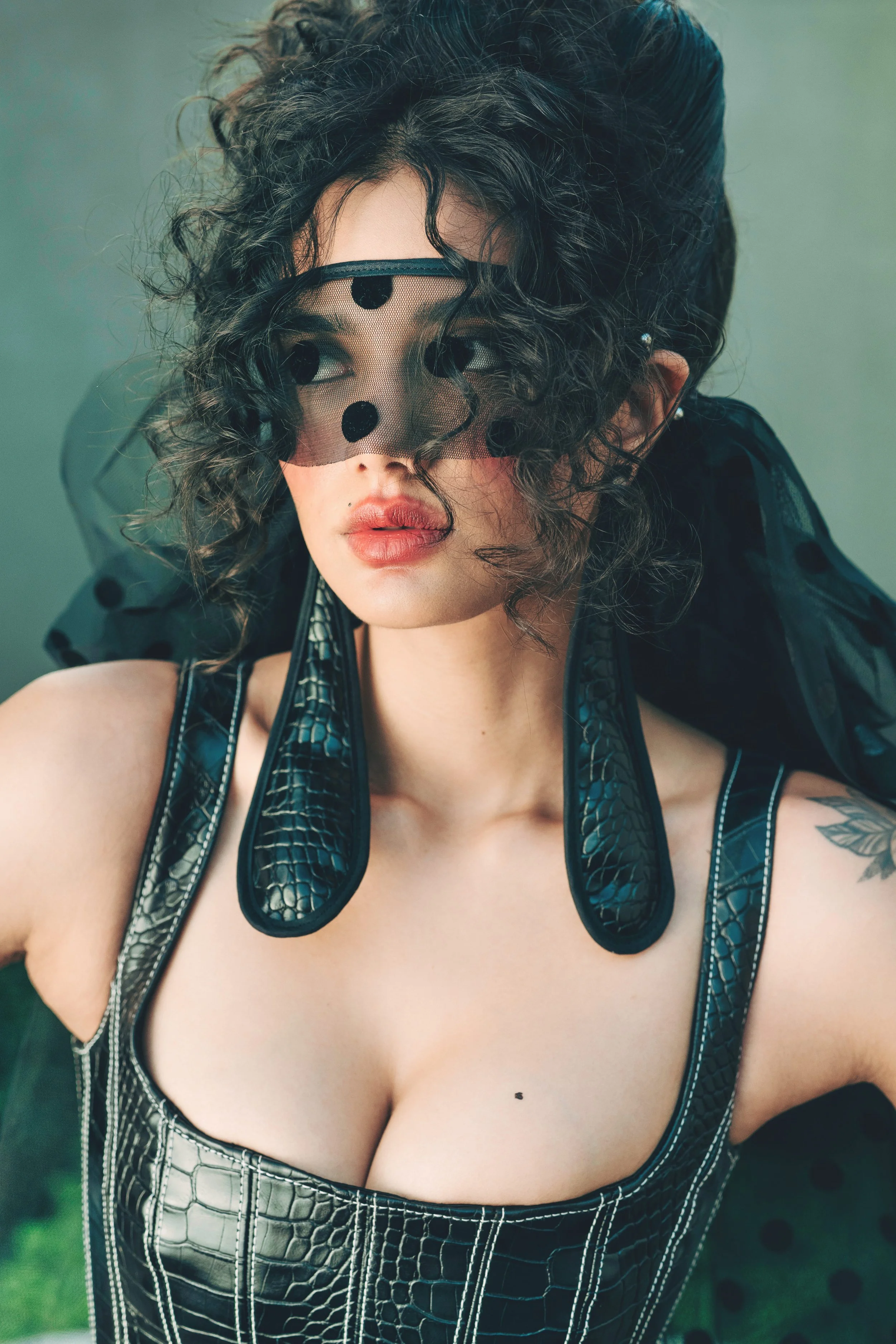 EMILY Francis from AURA Models with curly dark hair wearing a black mesh face mask with polka dots, a black leather top with a crocodile skin pattern, and earrings, posing indoors.