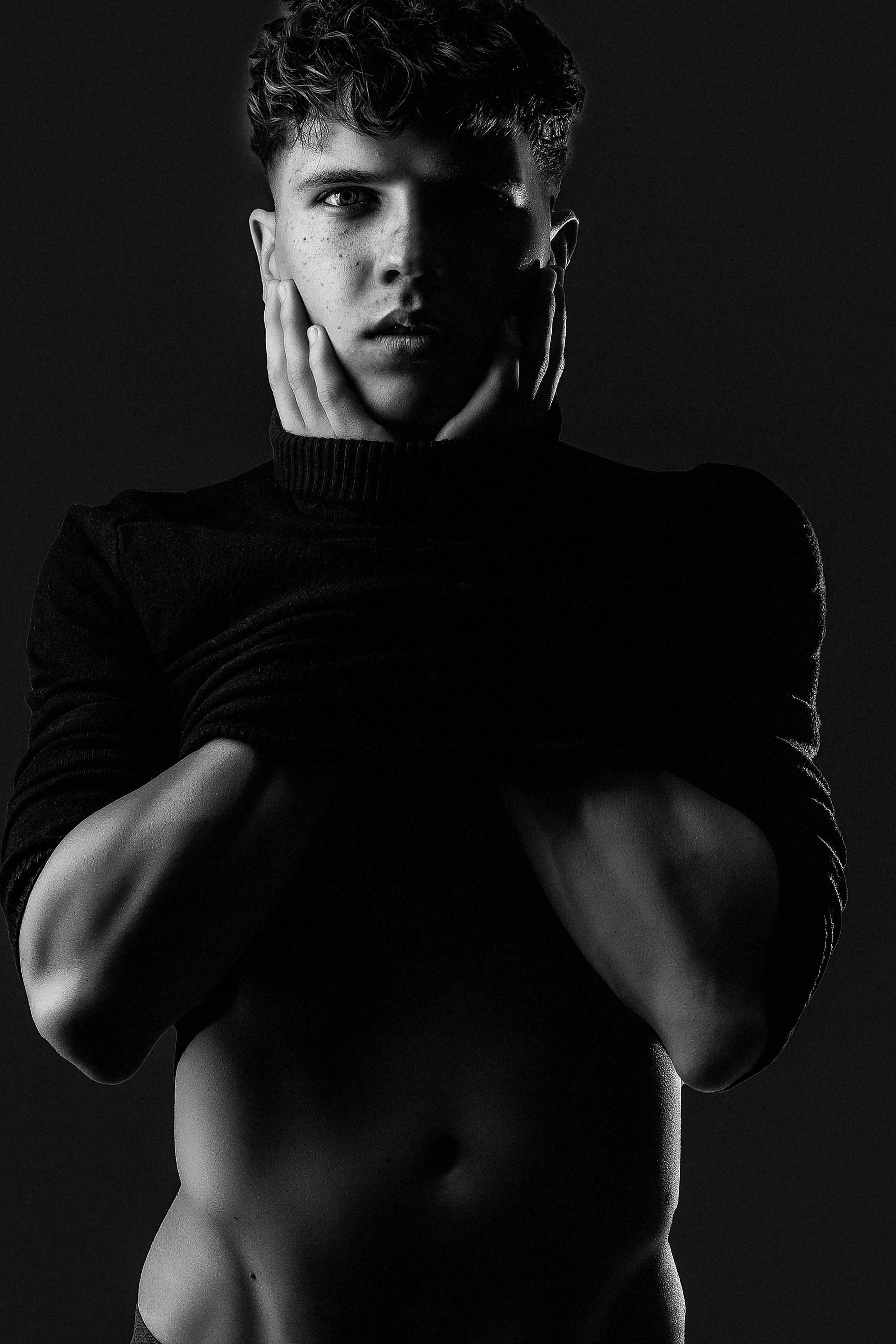Black and white portrait of a young man with short curly hair, shirtless, with hands on his cheeks, wearing a dark turtleneck sweater, against a dark background.