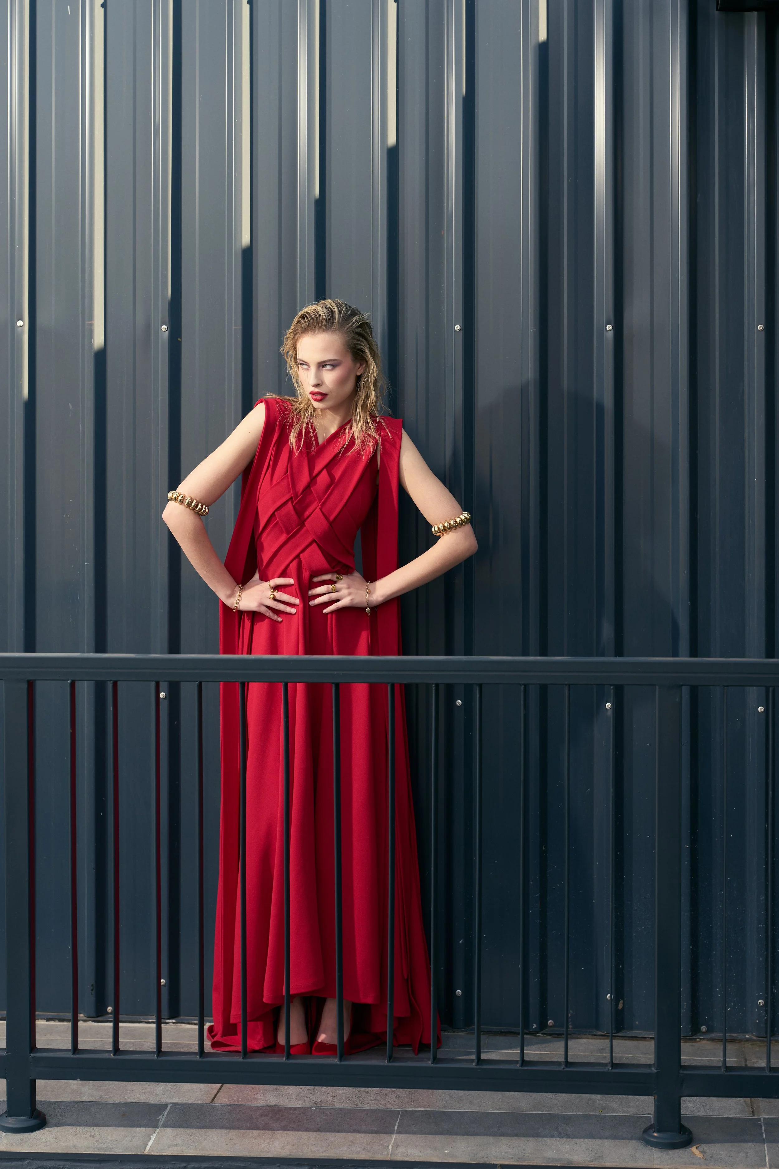 GAIA Fernandez from AURA Models wearing a red dress and gold jewelry standing behind a black metal fence with a blue corrugated metal wall in the background.