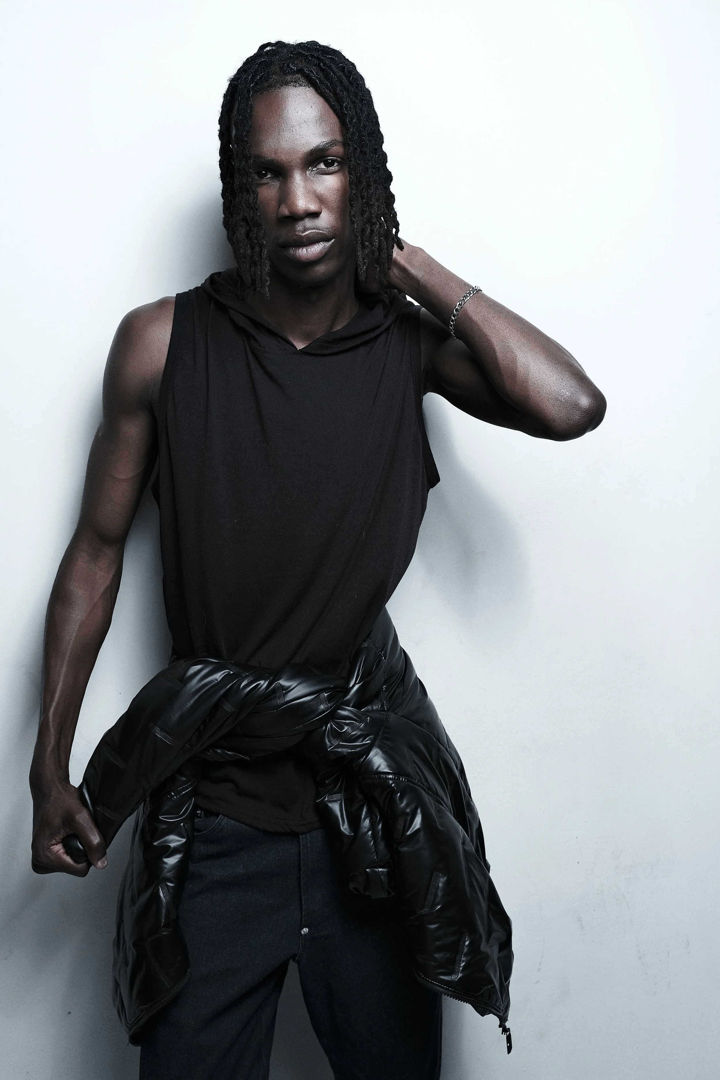 TSHEDZA Ramavheya from AURA with shoulder-length curly hair and dark skin, standing against a plain light-colored wall, wearing a sleeveless black shirt, dark jeans, and a black jacket tied around his waist, with his left hand resting behind his neck