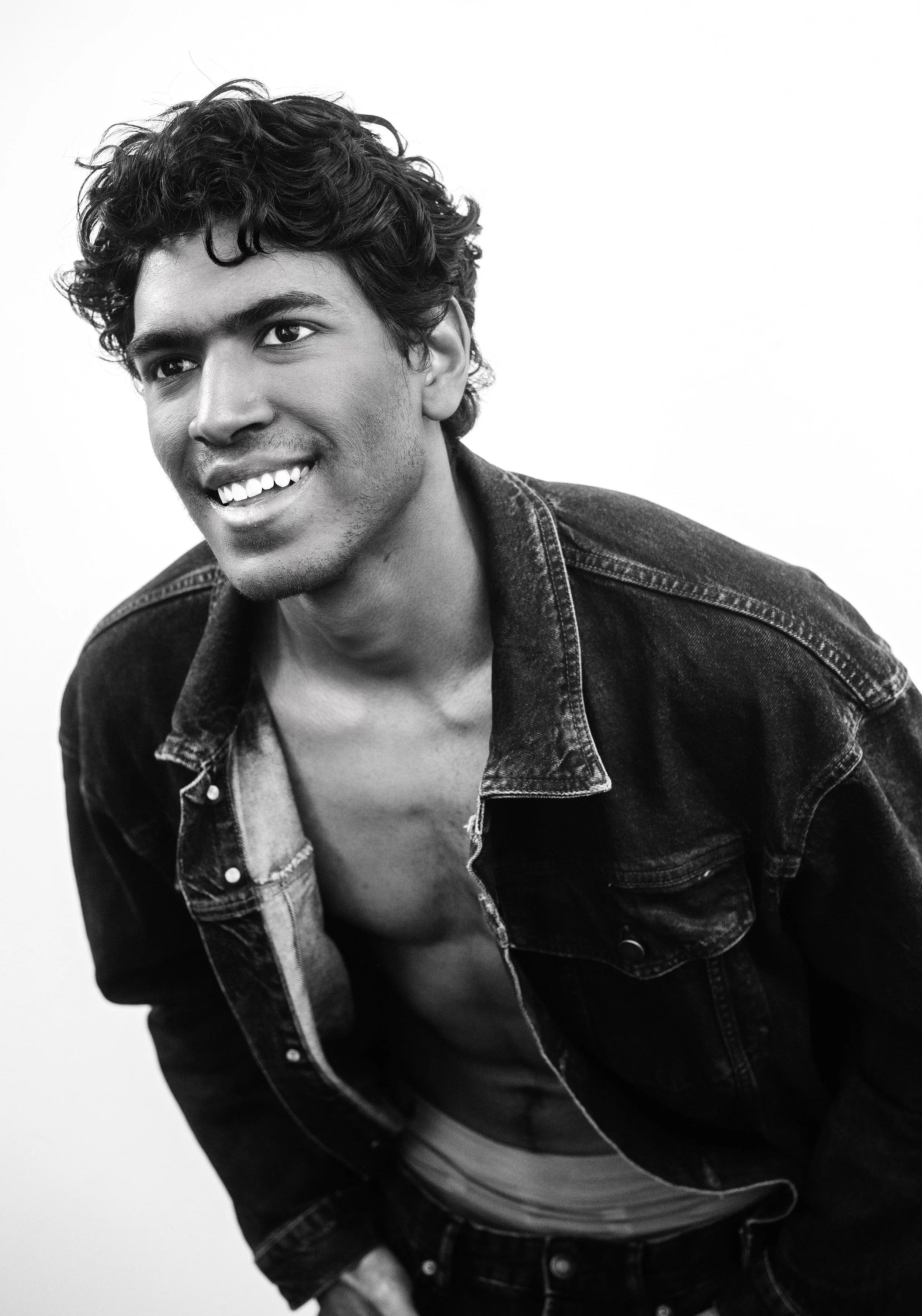 Black and white portrait of CALEB Naidoo from AURA Models with curly hair, smiling, wearing a denim jacket over a striped shirt, standing against a plain white background.