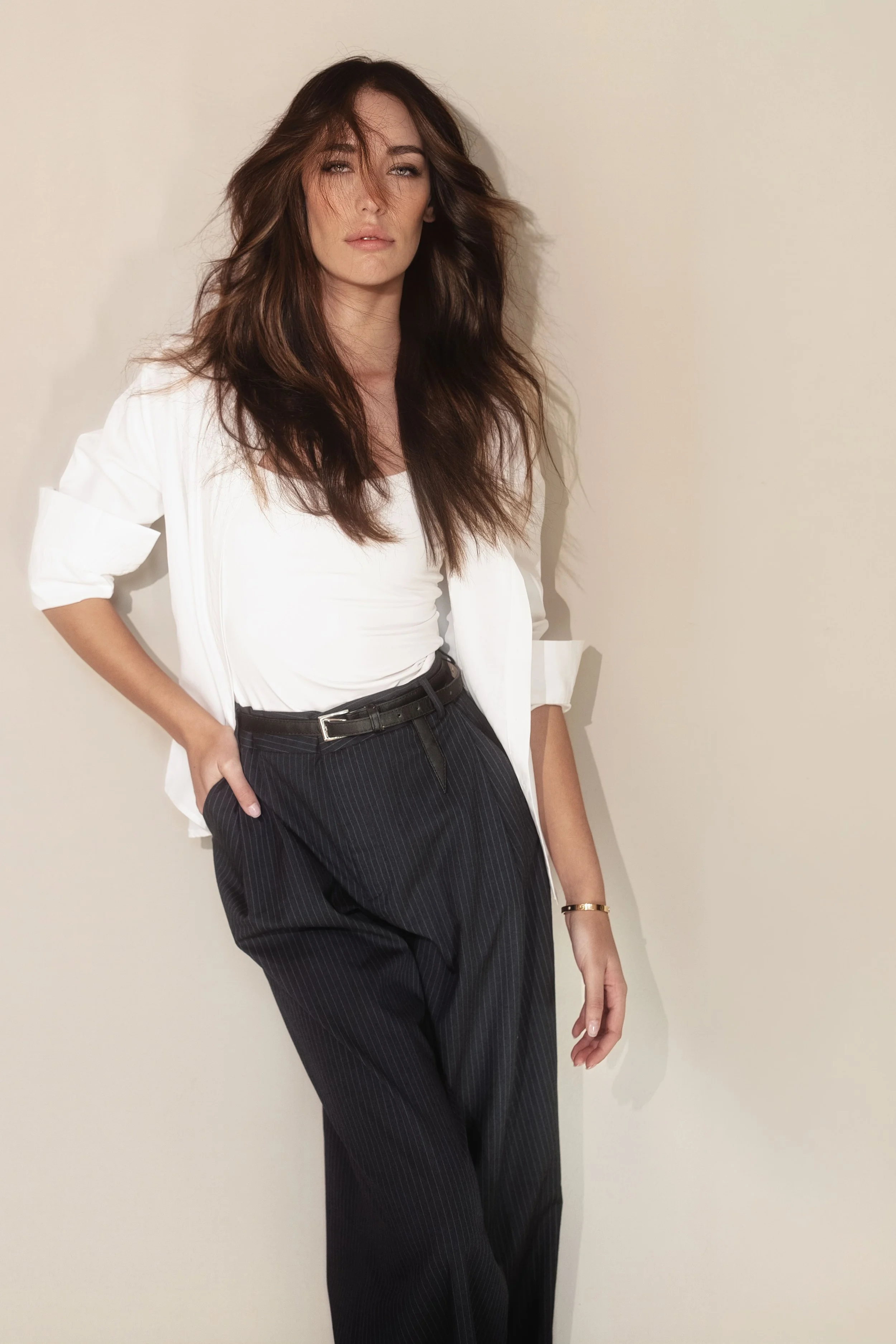 MONIQUE van Loggerenberg from AURA with long, wavy brown hair and light makeup, wearing a white blazer over a white top and high-waisted black pinstriped pants, standing against a plain off-white wall, with a relaxed pose and one hand in her pocket.