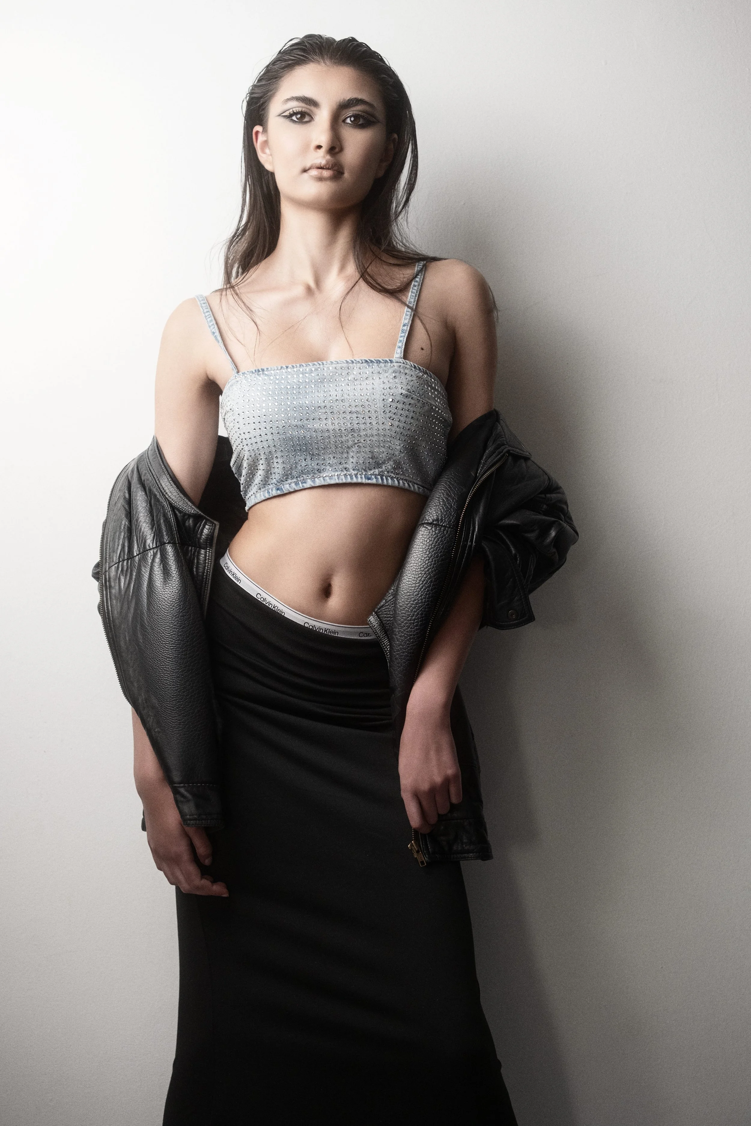 KEIRA Louw from AURA Models with long dark hair and makeup styling posing against a plain wall. She is wearing a sparkly crop top, a black skirt, and a black leather jacket draped off her shoulders.