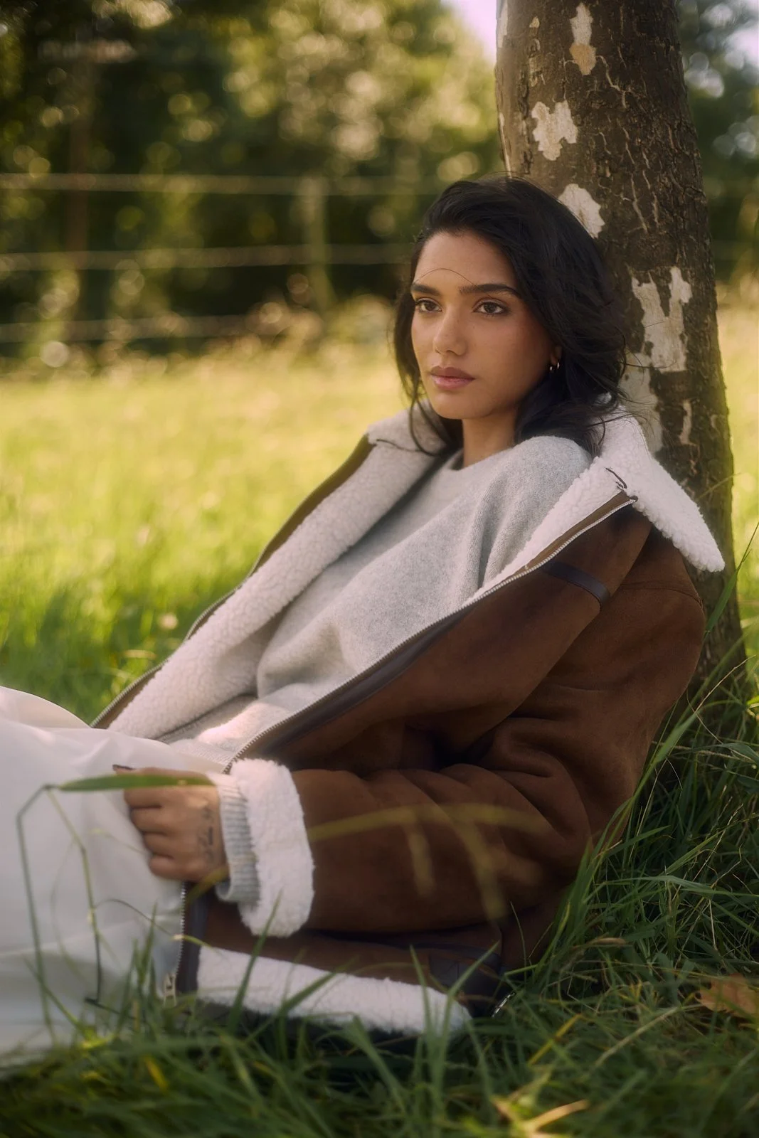 EMILY Francis from AURA Models with dark hair and light skin leaning against a tree outdoors, wearing a brown jacket with a sherpa lining and a gray sweatshirt, in a grassy area with blurred trees and sunlight in the background.