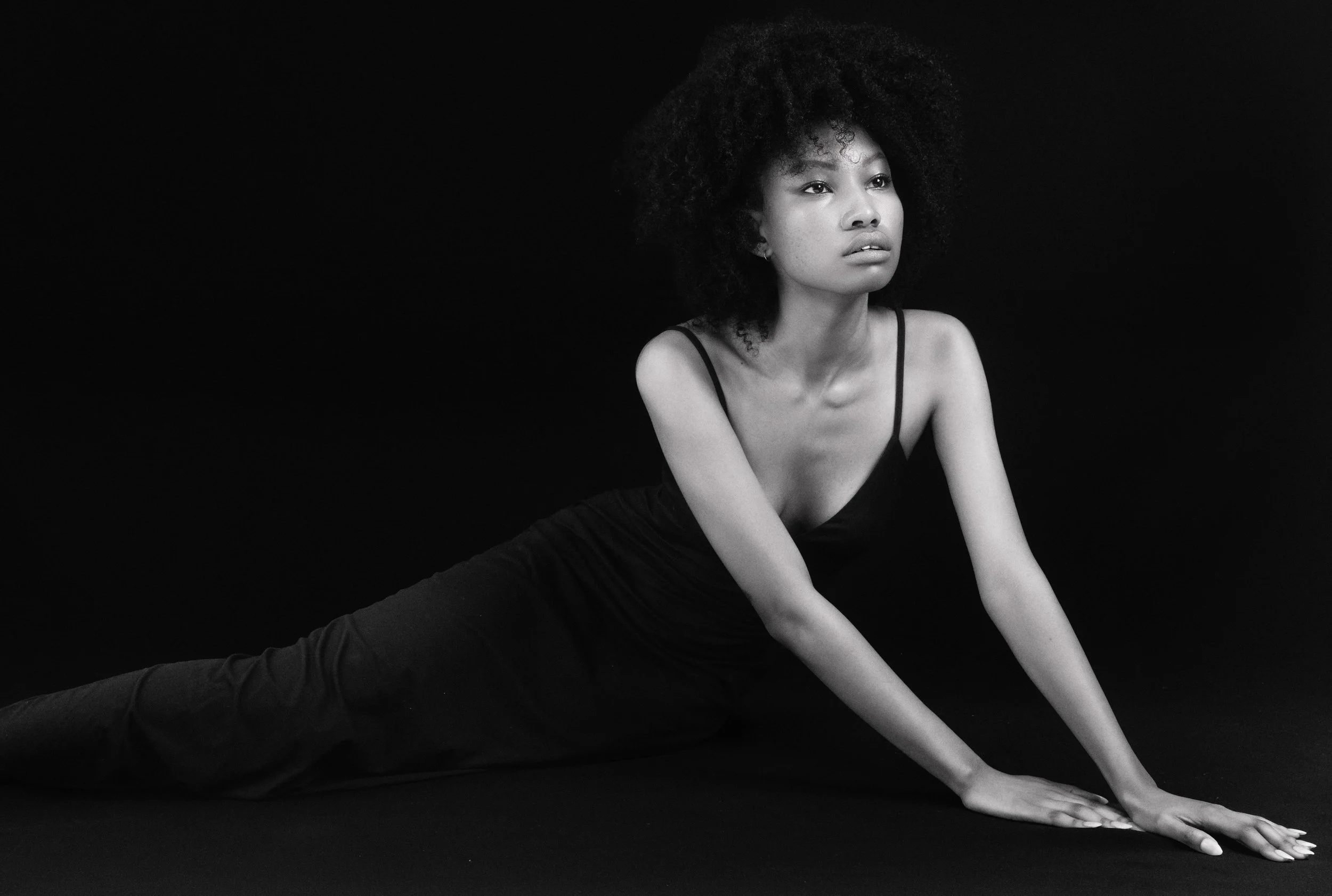 QUISHA Rapoo from AURA Models with curly hair in a black dress stretching on the floor against a black background.