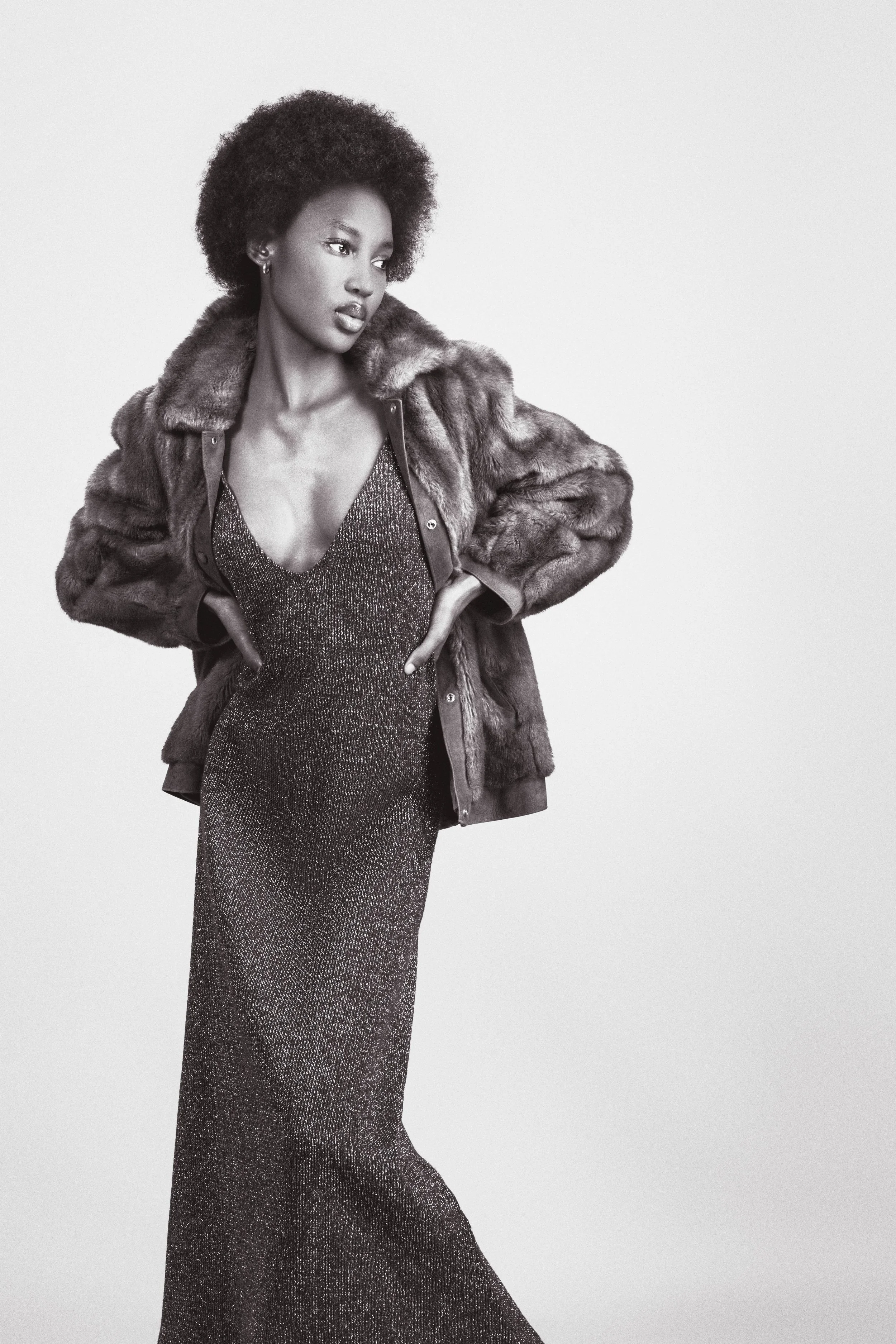 Black and white photo of RORY Merafe from AURA Models wearing a knee-length sparkly dress and a fur jacket, posing with her hands on her hips, looking to the side.