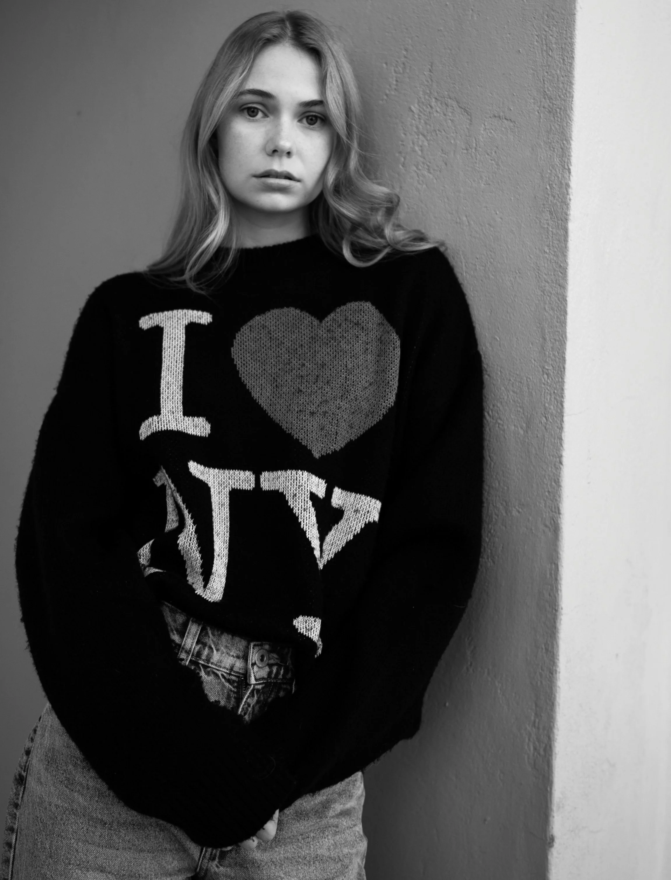 A young woman with wavy hair, standing against a textured wall, wearing a black sweater with a heart and the words 'I love NY', and light-colored jeans.