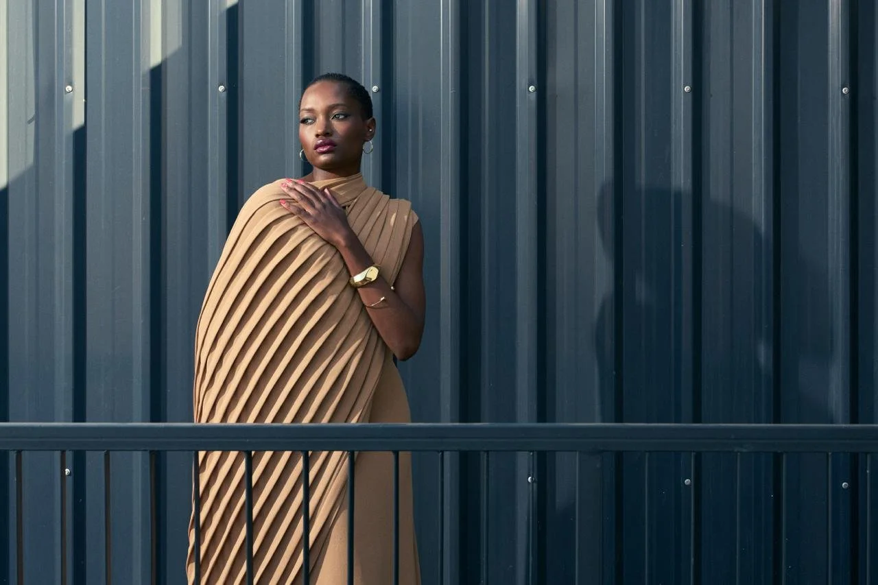 SHALOM Musvosvi from AURA Models, wearing a tan pleated dress, standing in front of a dark gray metal fence.