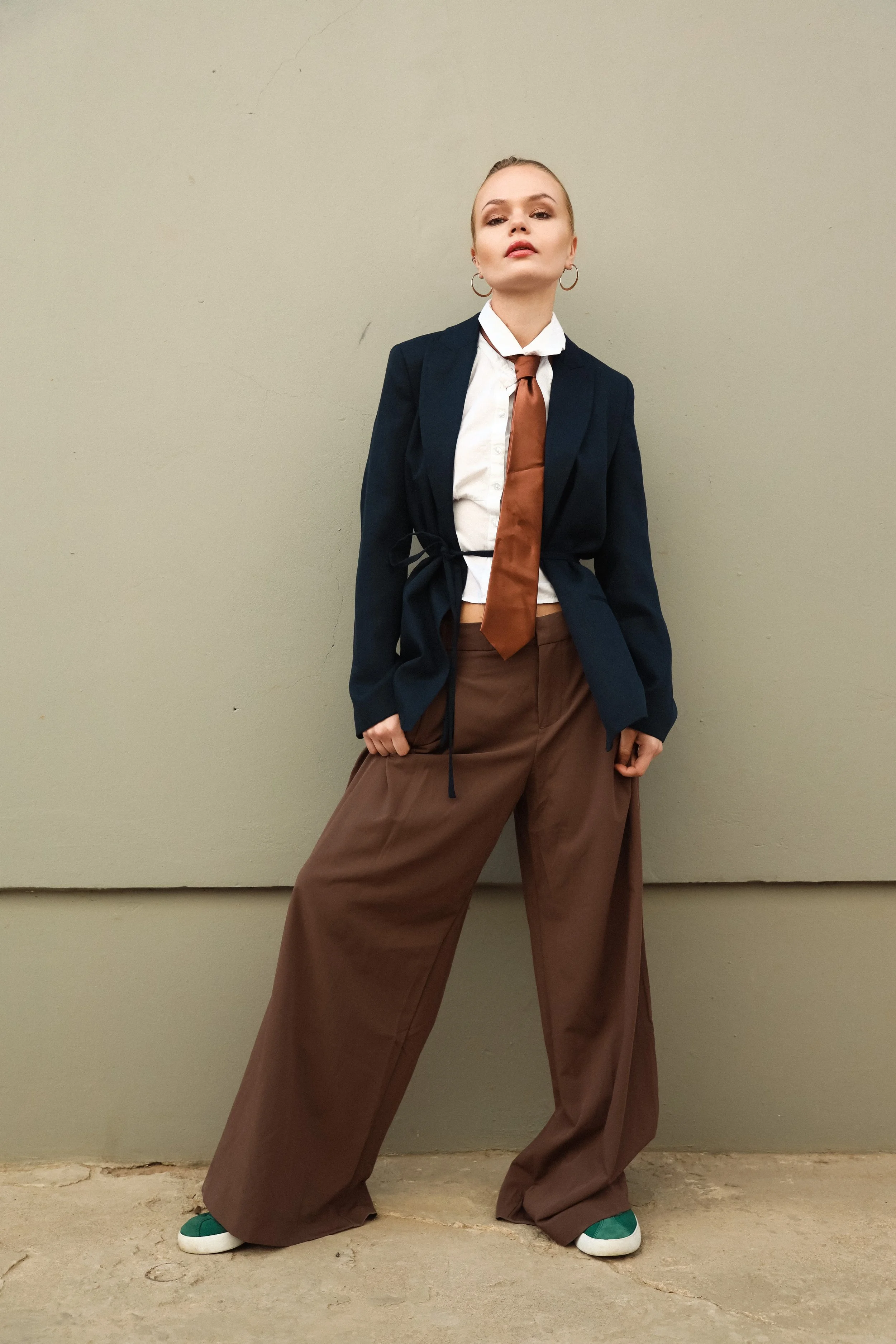 A person with blonde hair styled in a tight bun, wearing makeup, in a white shirt, brown wide-leg pants, a navy blazer, and a brown tie. They stand against a plain wall, with one hand in their pocket and the other hanging down, wearing green and whit