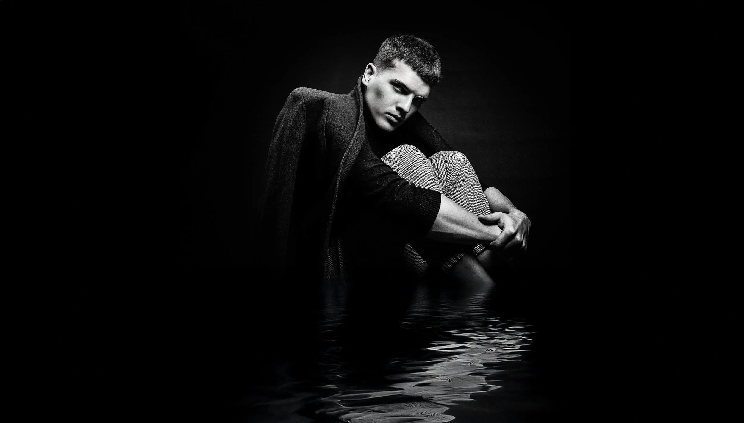 Black and white photo of a person sitting with knees drawn up, wearing a coat and patterned pants, in water with ripples, against a dark background.