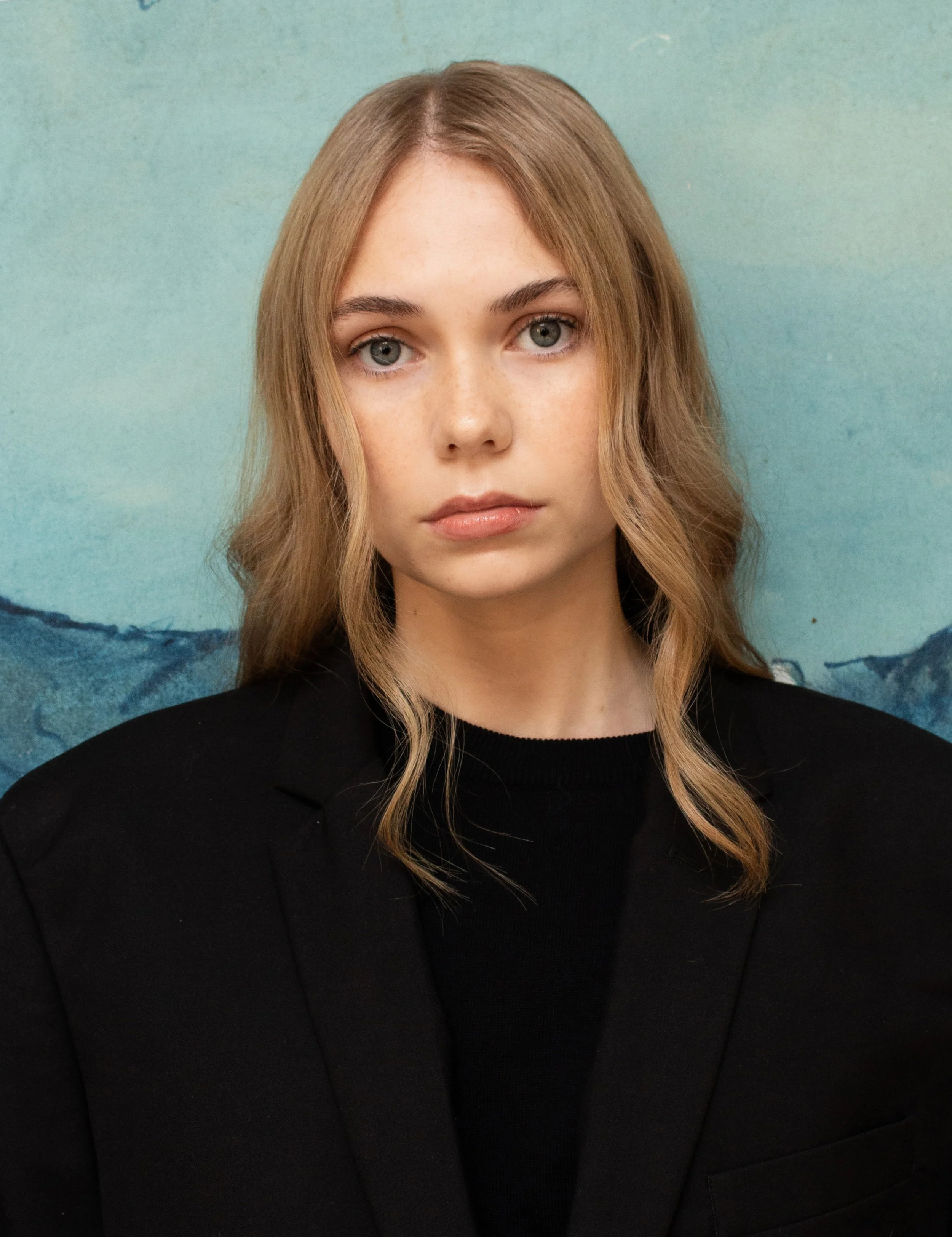 A woman with long wavy blonde hair, wearing a black blazer, looking directly at the camera against a blue-green background.