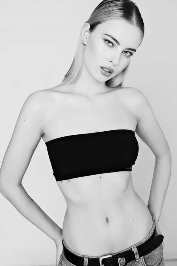 Black and white photo of GAIA Fernandez from AURA Models  with light hair, wearing a black strapless top and jeans, posing with one hand on her hip.