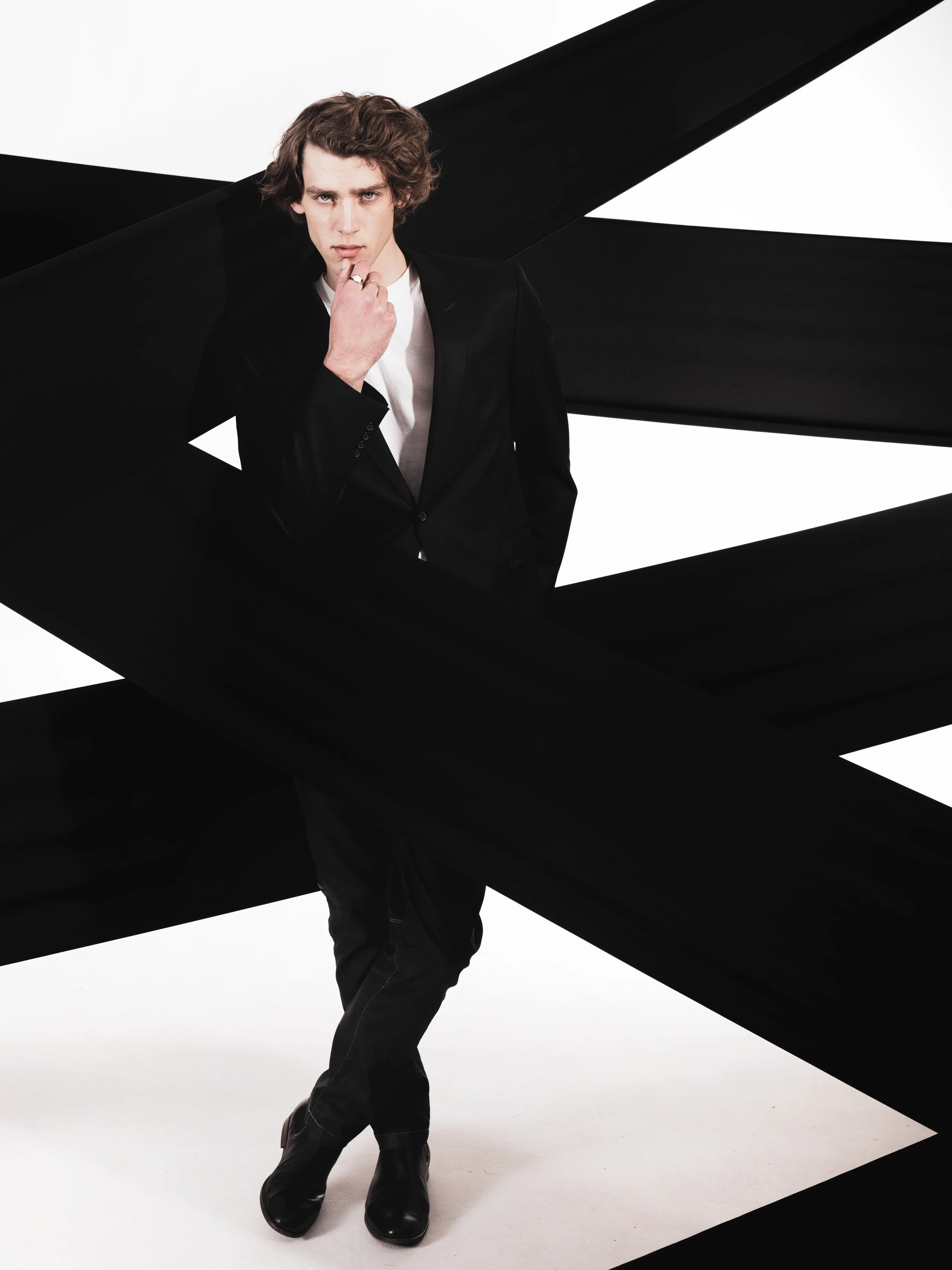 JOSH Gradwell from AURA Model with curly brown hair, wearing a black jacket, white shirt, and black pants, standing in front of a black and white abstract background with geometric shapes, looking directly at the camera with one hand near his chin.