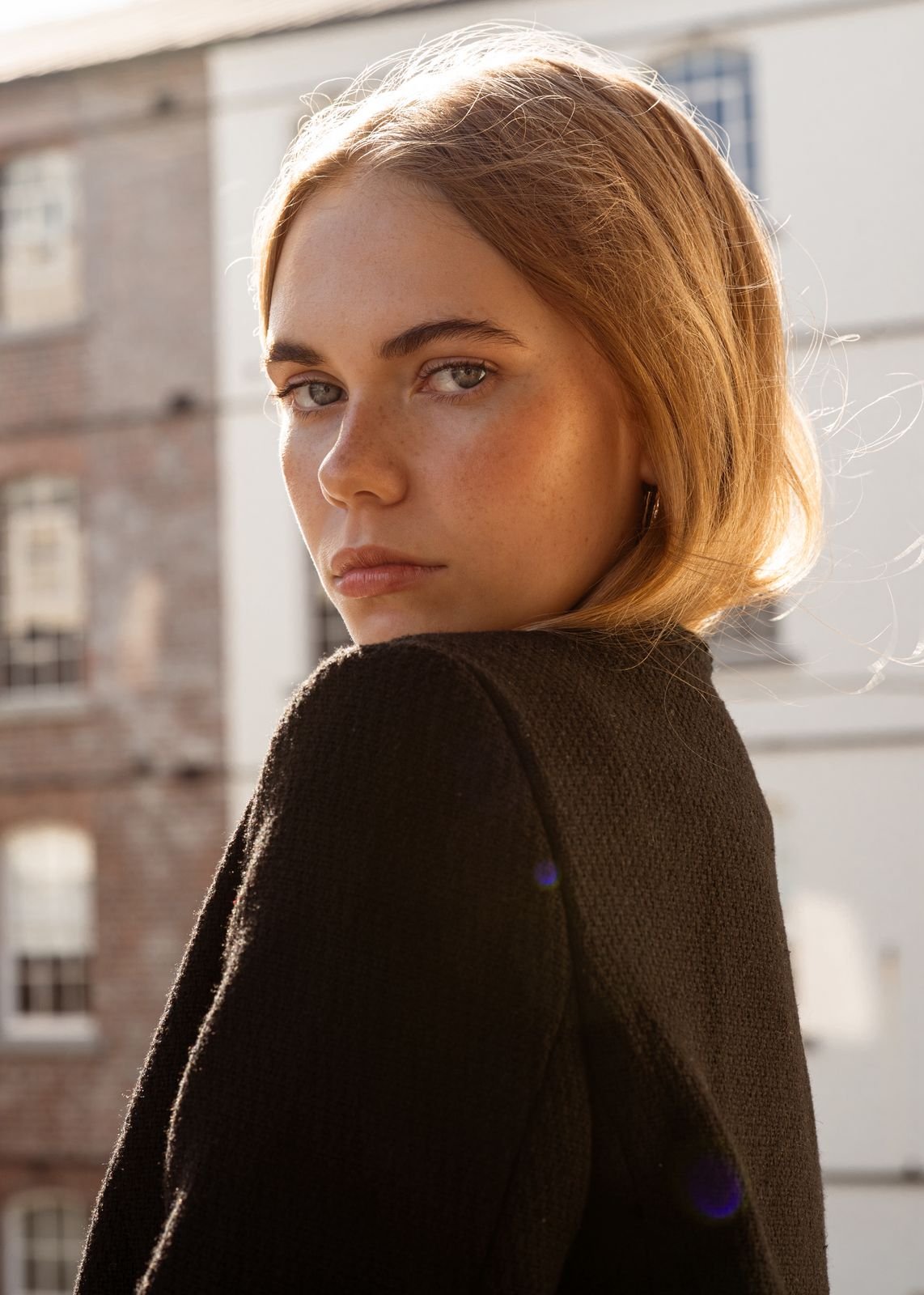 KEELY Miller from AURA Models with red hair and blue eyes looking over her shoulder outdoors, with sunlight illuminating her face and a blurred cityscape background.