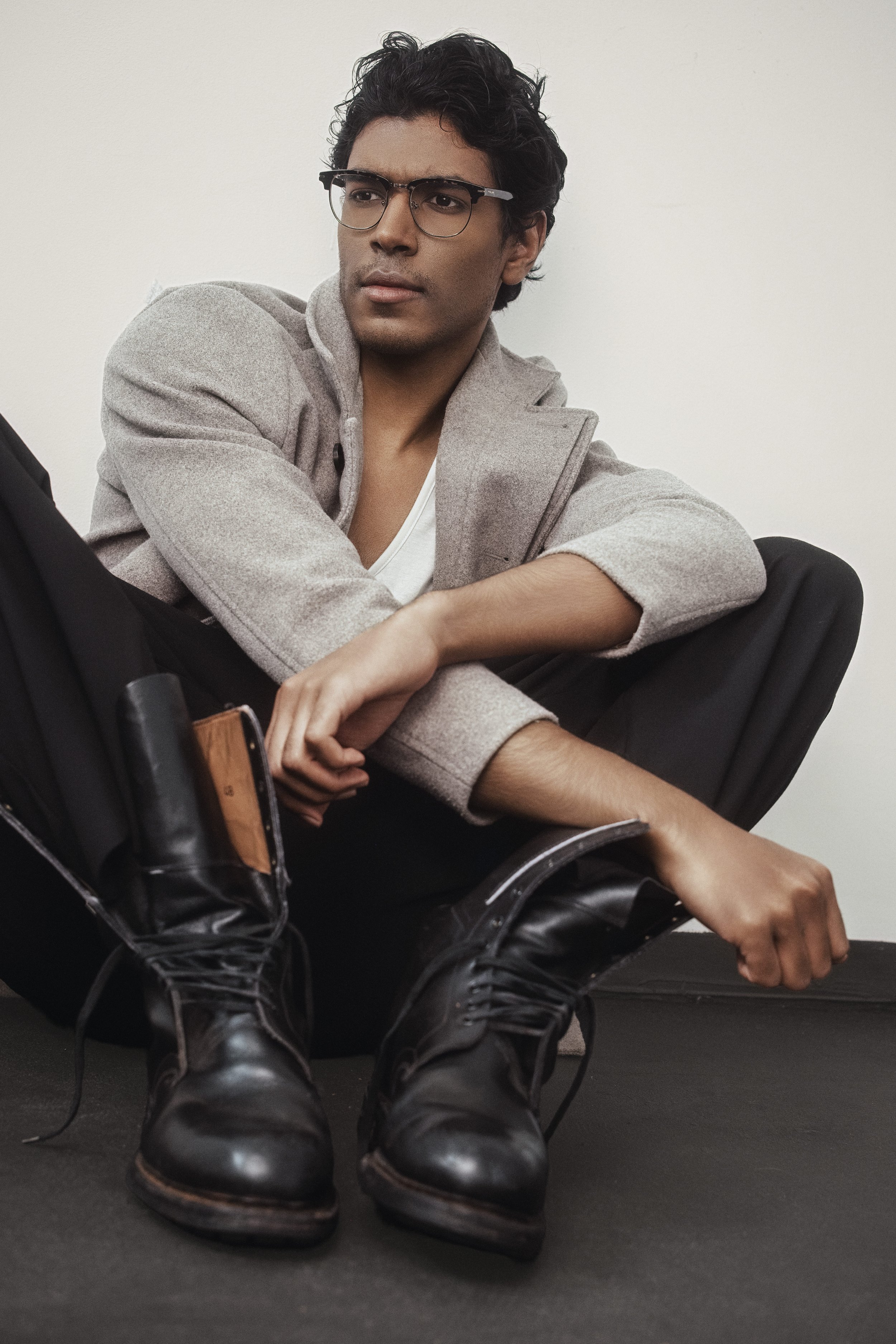 CALEB Naidoo from AURA Models sitting on the floor with crossing legs, wearing glasses, a gray blazer, white shirt, black boots, and holding a backpack.