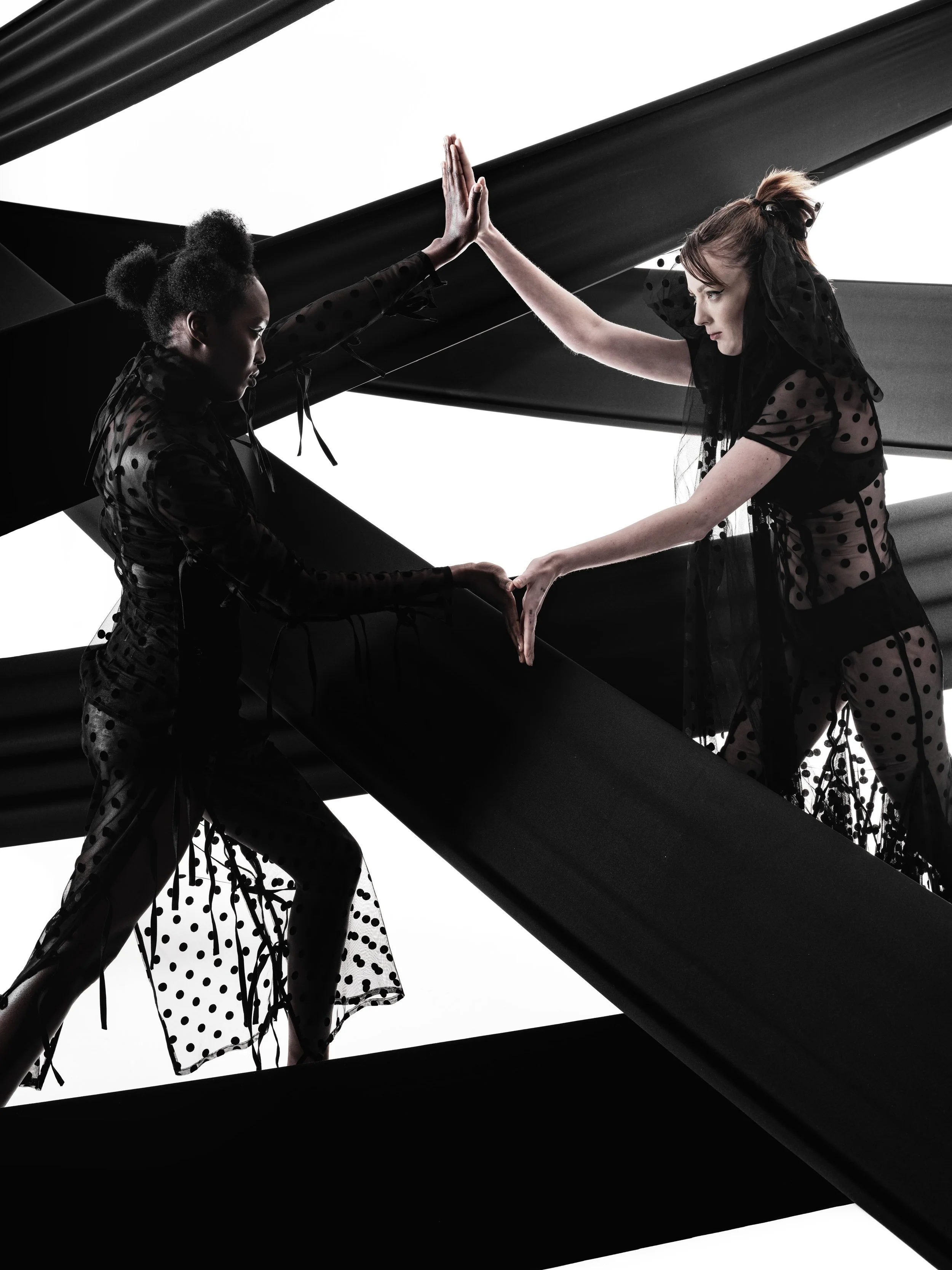 CAYLEIGH Bancroft and KIM Chakanyuka from AURA Models dressed in sheer black polka dot clothing are playing a high-energy game of ping-pong, with one woman about to hit the ball and the other about to return it.
