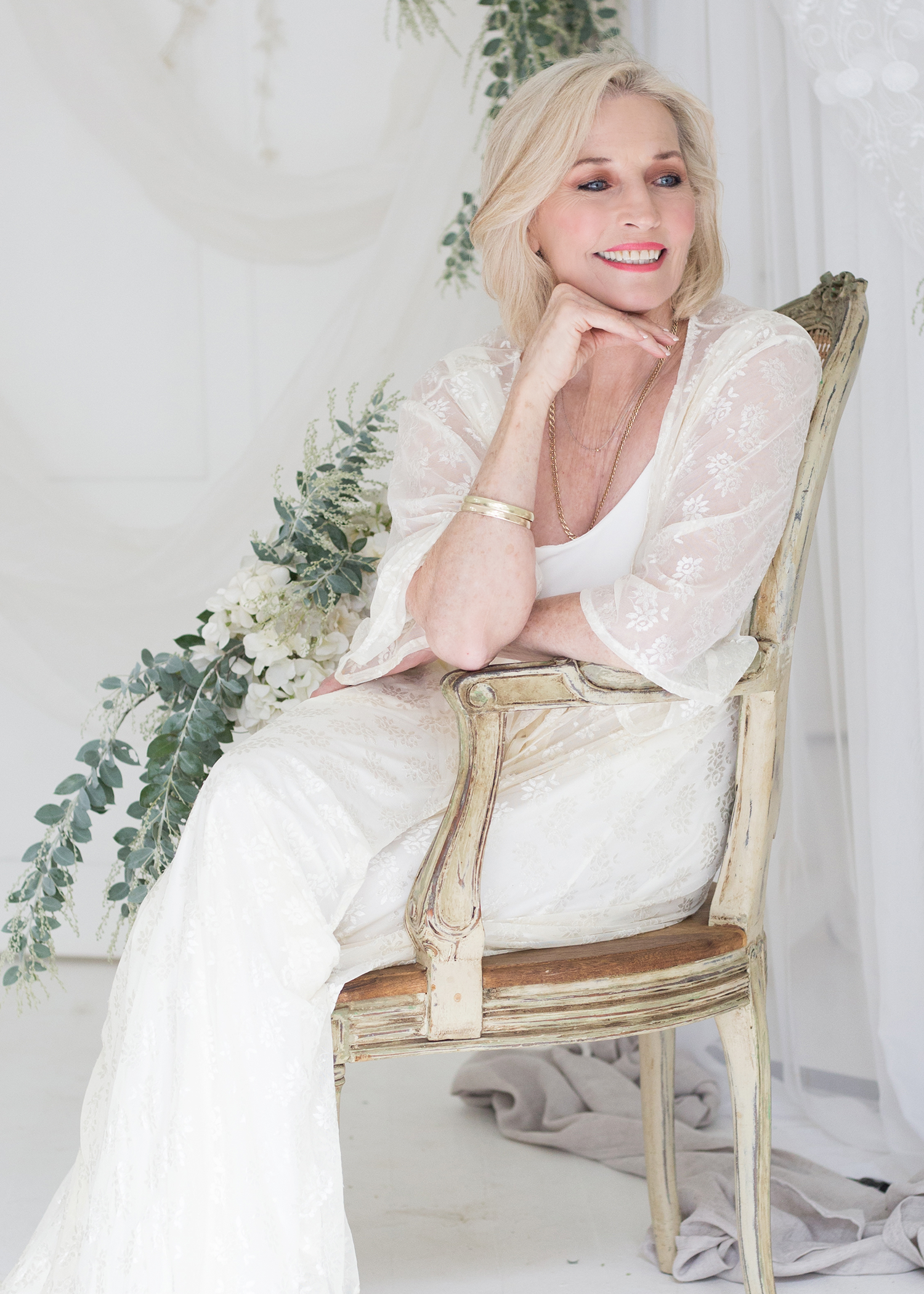 MARIETA Ferreira from AURA Models with blonde hair sitting on a wooden antique chair, wearing a white lace crochet dress, and smiling while looking to her right in a light, airy room decorated with green foliage and white flowers.