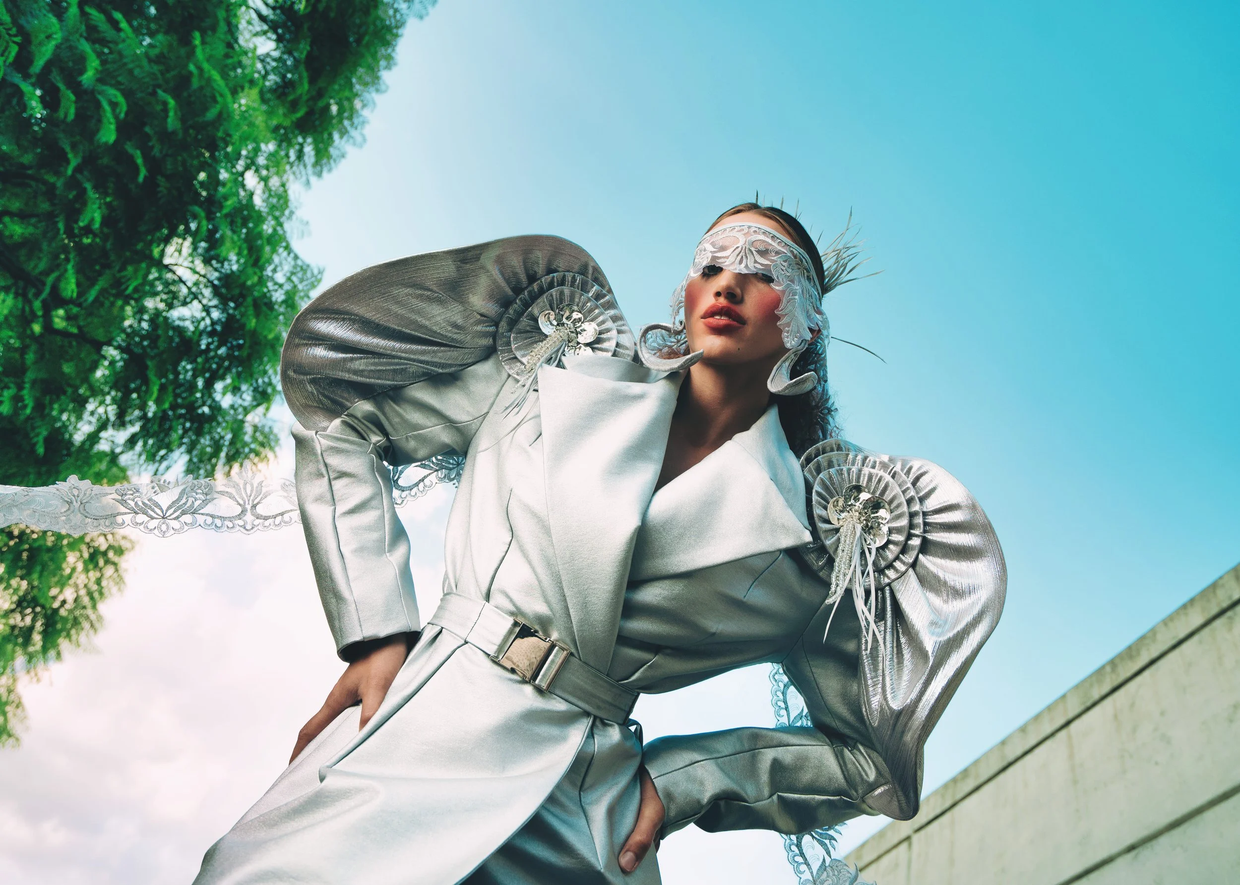 MEGAN Leigh from AURA Models dressed in a futuristic silver outfit with large, artistic shoulder accents, standing outdoors against a backdrop of green foliage and a bright blue sky. She is blindfolded with lace and has feathers in her hair.