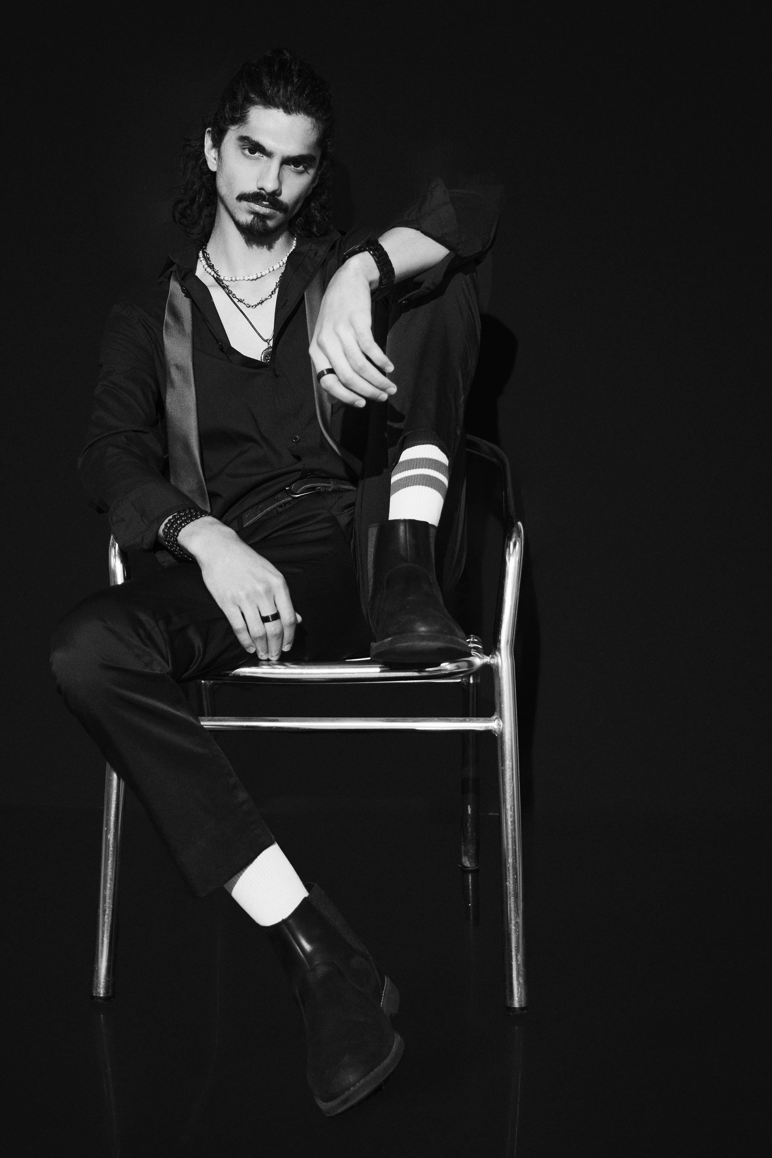 Black and white portrait of a man with a mustache and goatee, sitting on a metal chair. He is wearing dark clothing, a necklace, rings, bracelets, and black boots. He has long curly hair and is posed with one leg crossed over the other, with an inten