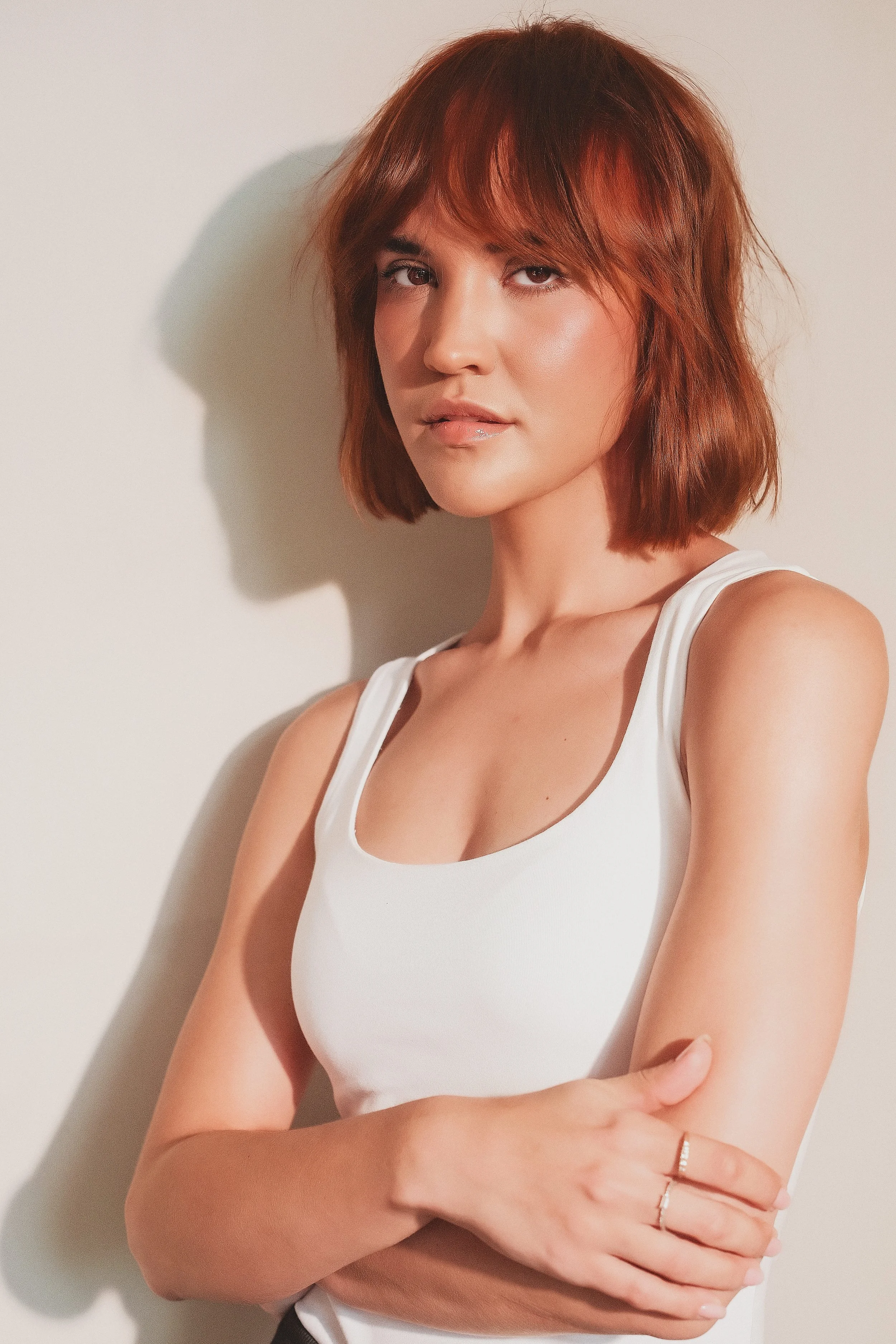 MARINET Potgieter from AURA Models Urban with shoulder-length reddish-brown hair wearing a white tank top, standing against a plain light-colored wall, with one arm crossed over her body and a neutral expression.