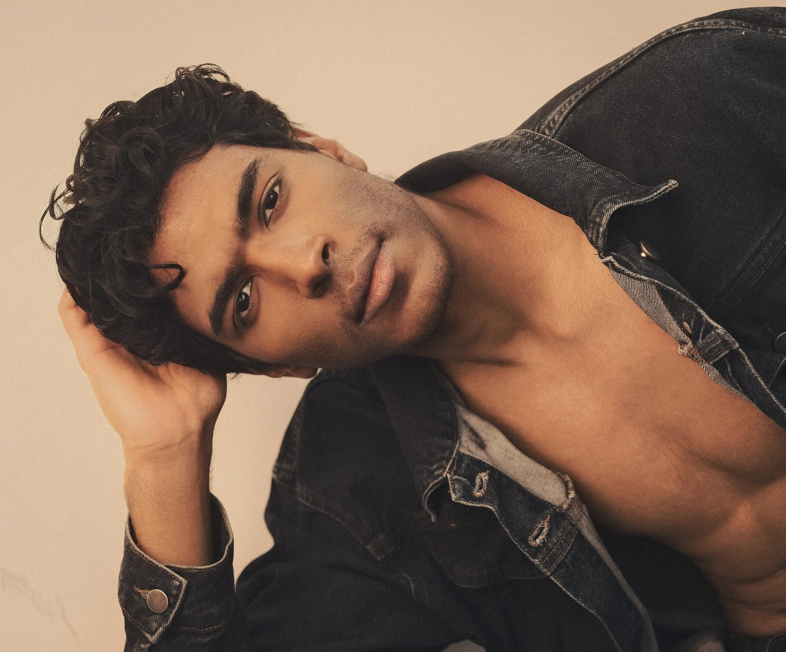 CALEB Naidoo from AURA Models with dark, curly hair and a warm smile, lying on his side with his head resting on one hand, wearing a black denim jacket, against a neutral background.