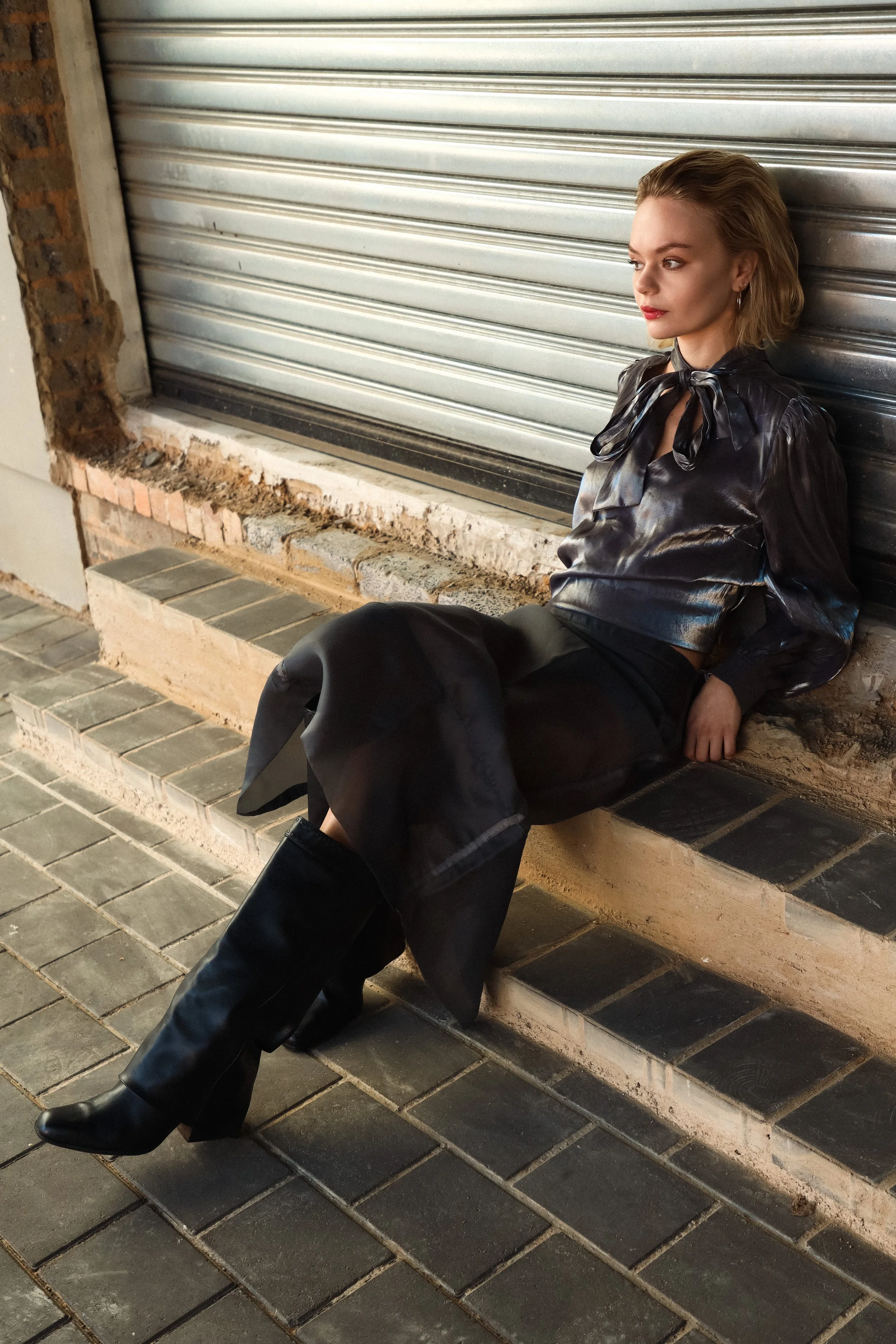 A young woman with blonde hair and red lipstick sitting on a concrete ledge outside, leaning against a metallic roll-up door, wearing a black leather blouse with a bow tie at the collar, black trousers, and black boots.