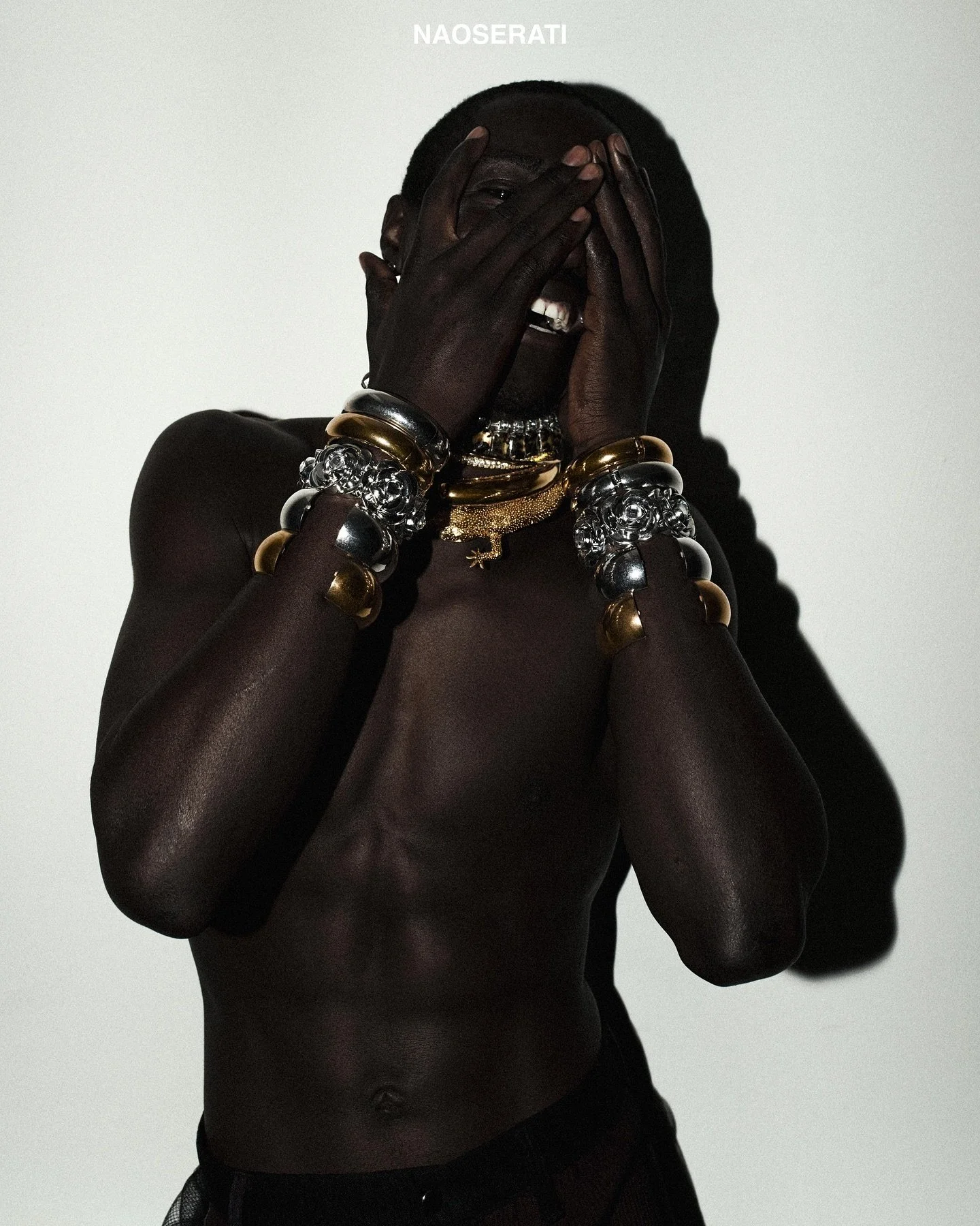 A shirtless man with dark skin covering part of his face with his hands, wearing multiple metallic and gold bracelets, a chain necklace, and smiling.