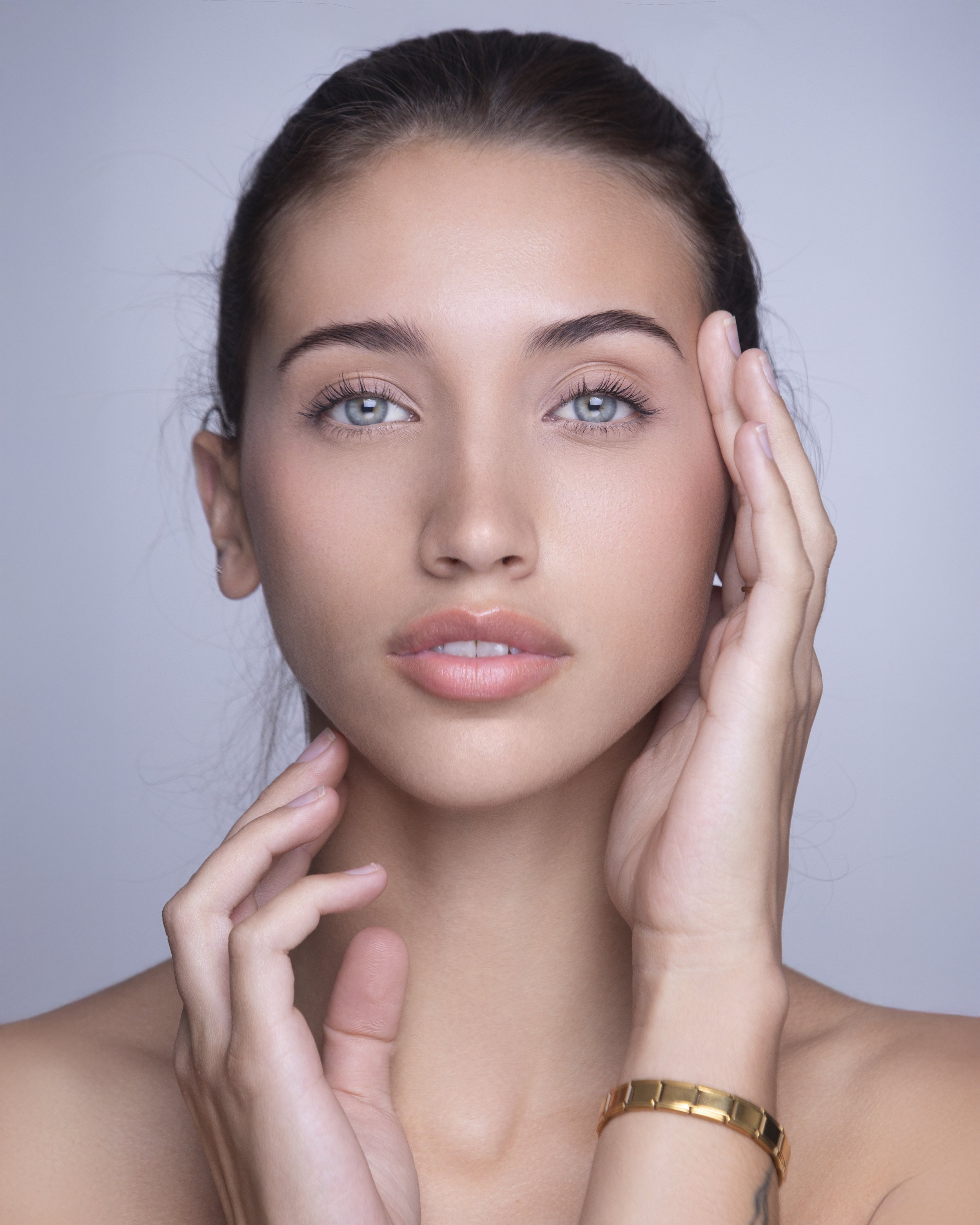 Close-up of a young woman with blue eyes, natural makeup, and dark hair pulled back, touching her face gently.