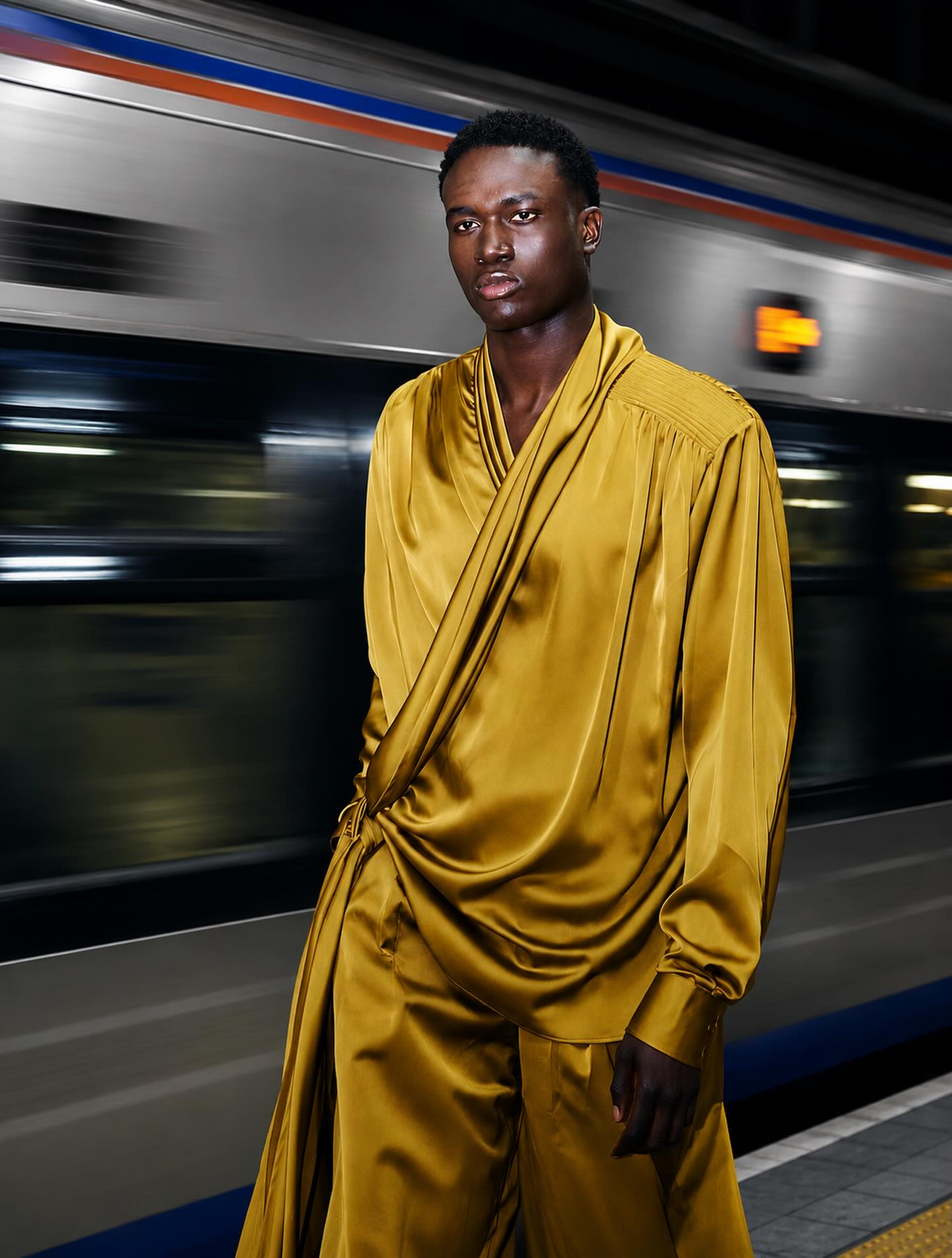 MBHURI Baloyi from AURA Models in a gold satin outfit standing on a subway platform with a moving train in the background.