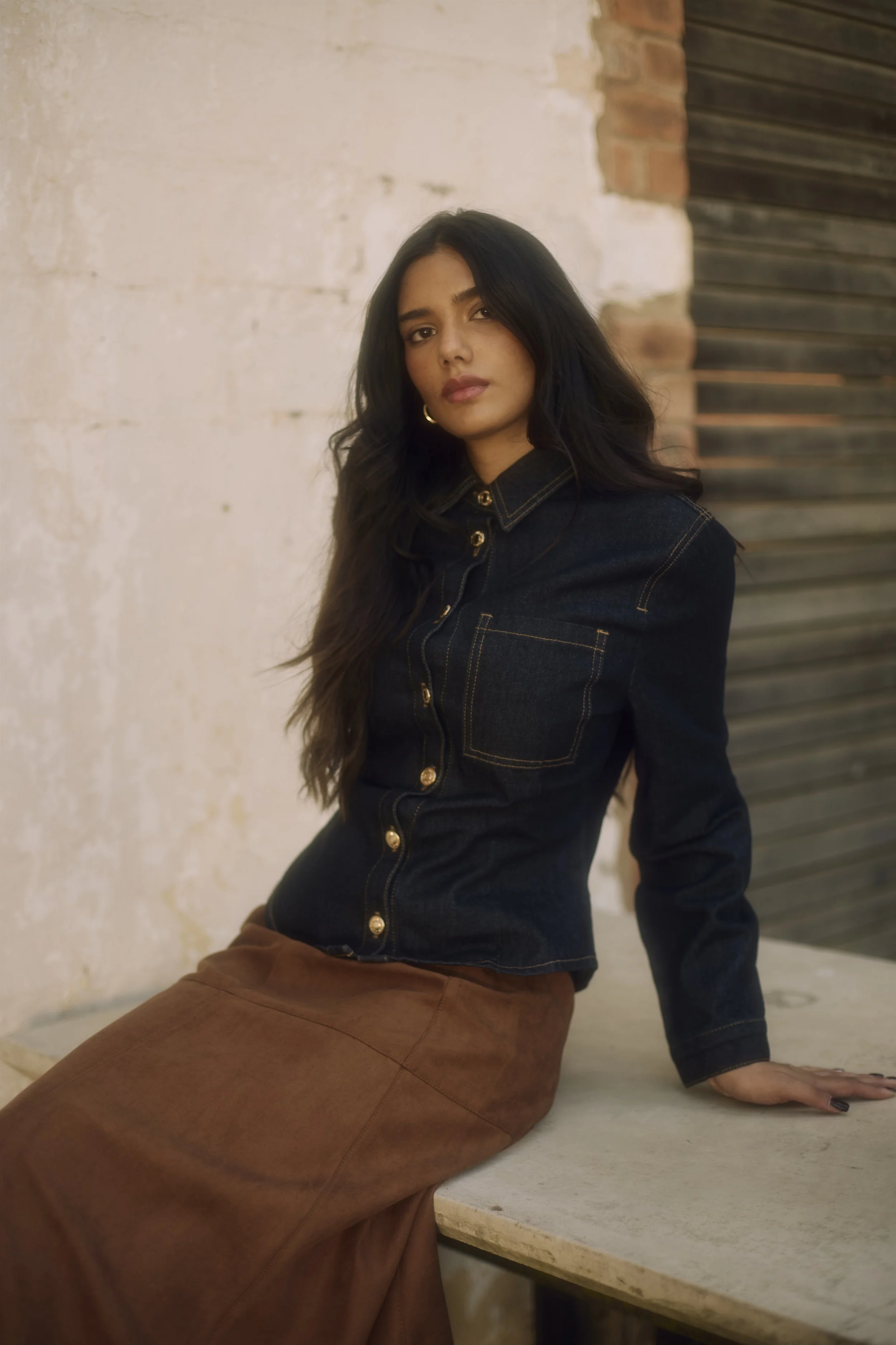 EMILY Francis from AURA Models with long dark hair wearing a dark denim jacket and brown skirt sitting on a marble surface against a rustic wall with brick and wooden shutter.