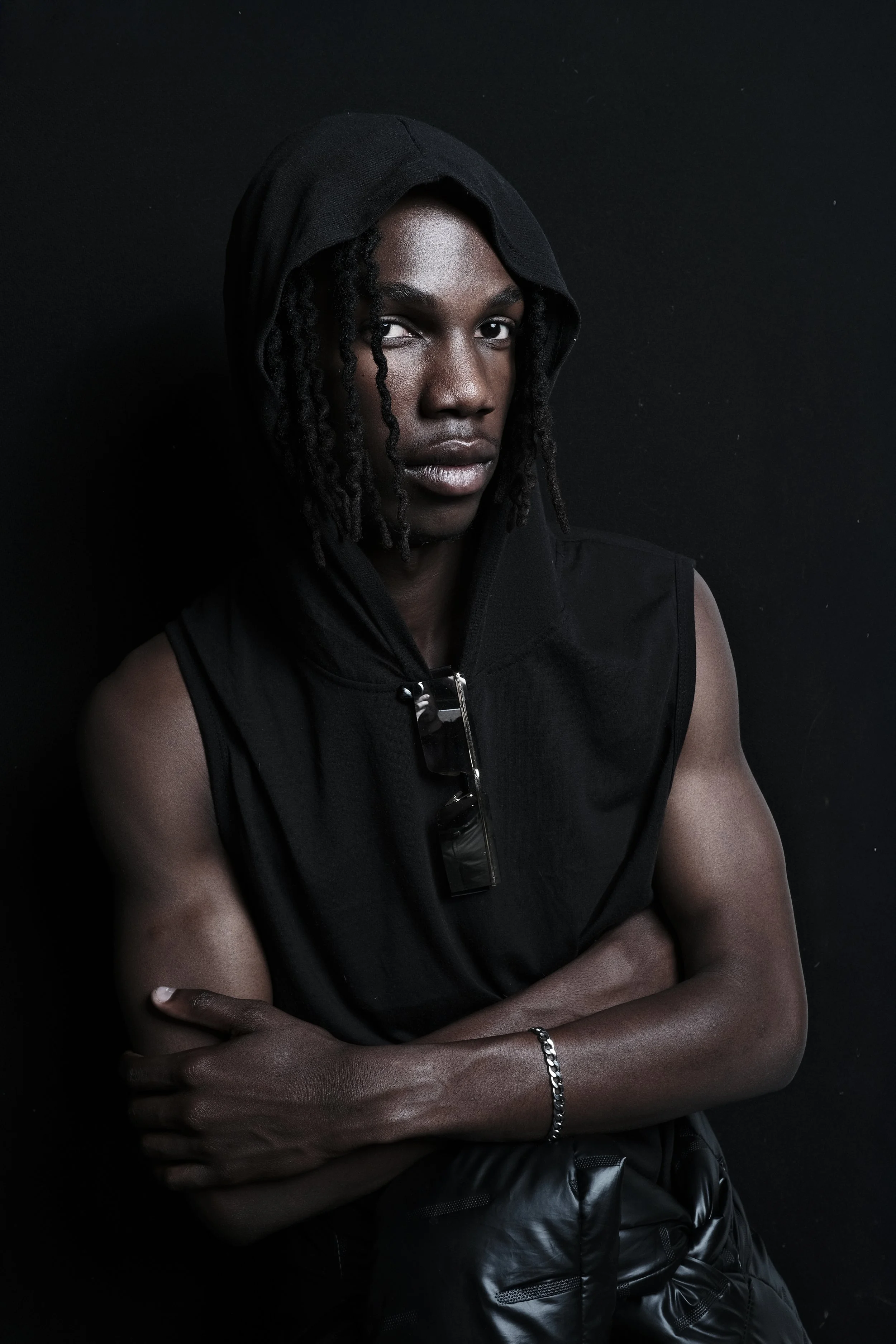 TSHEDZA Ramavheya from AURA Models with dark skin, dreadlocks, wearing a black sleeveless hoodie with the hood up, and a silver bracelet, standing against a black background.