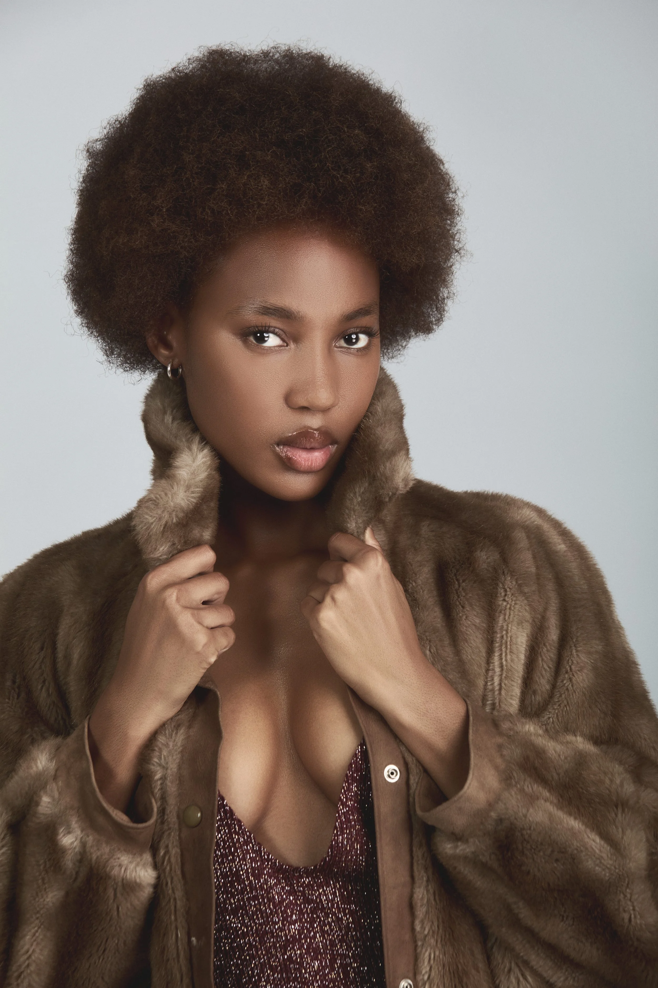 RORY Merafe from AURA Models with natural hair in a voluminous afro style, wearing a brown fur coat and a sparkling top, holds the coat collar with both hands, posing against a neutral background.